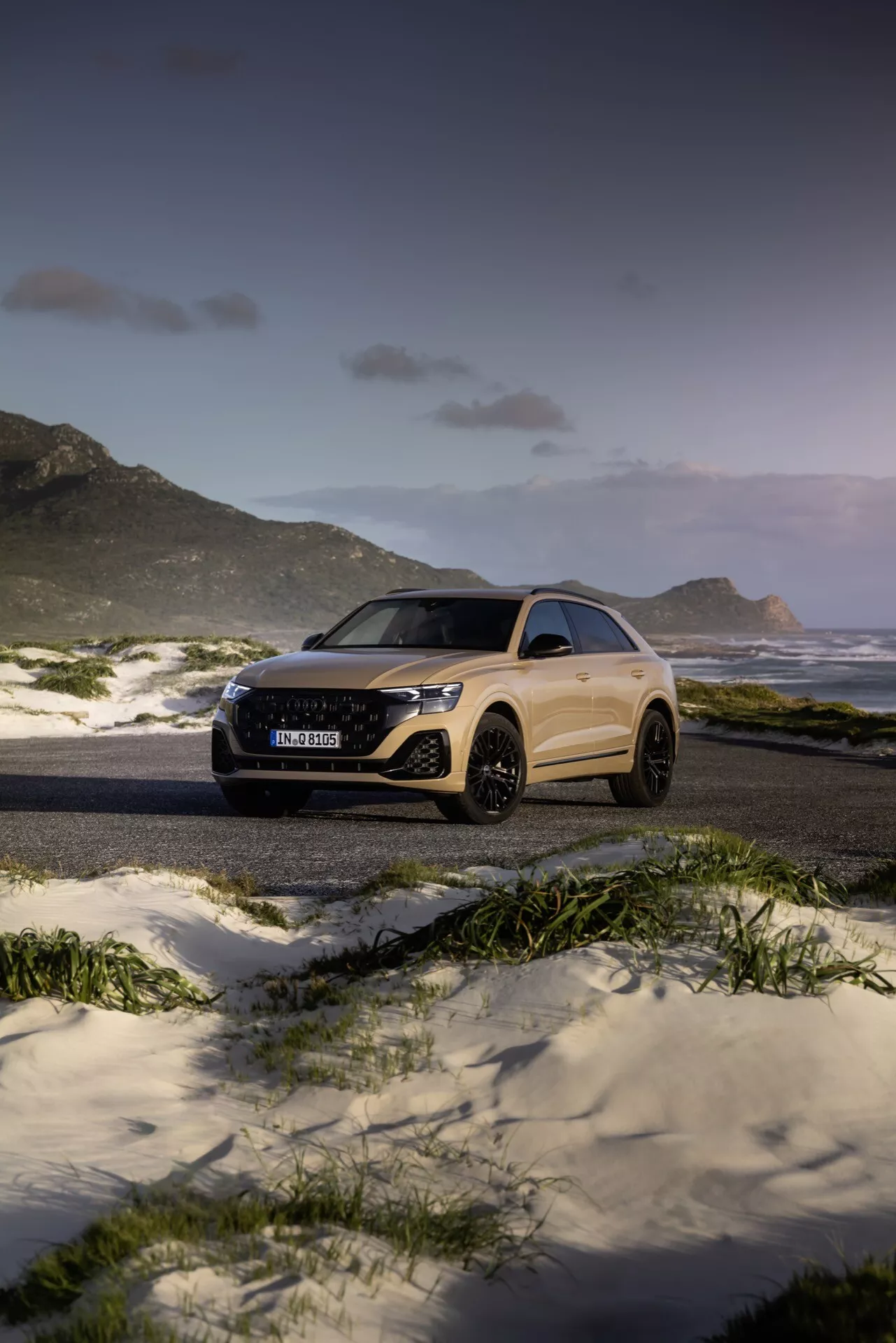 Audi Q8 2023 (4M-Facelifting) i ett majestätiskt strand- och bergslandskap i skymningen, som visar upp sin eleganta design och avancerade funktioner som LED-strålkastare och stora legeringshjul, från en tre fjärdedels fram- och sidovinkel