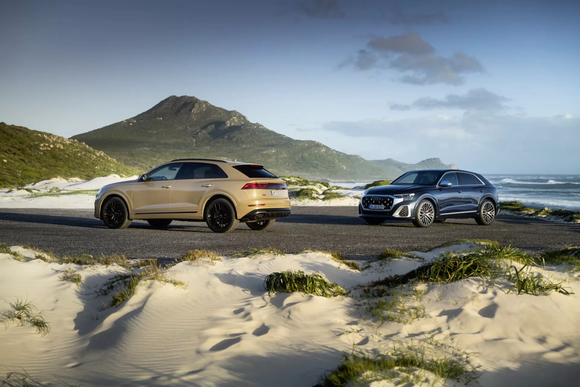 2023 års Audi Q8 (4M-facelift) modeller visas på en sandig strand, med en guld sport SUV till vänster i tre fjärdedelars bakre vy och en traditionell silver SUV till höger i tre fjärdedelars framsida vy, mot bakgrund av gräsbevuxna dyner, berg och en delvis molnig himmel