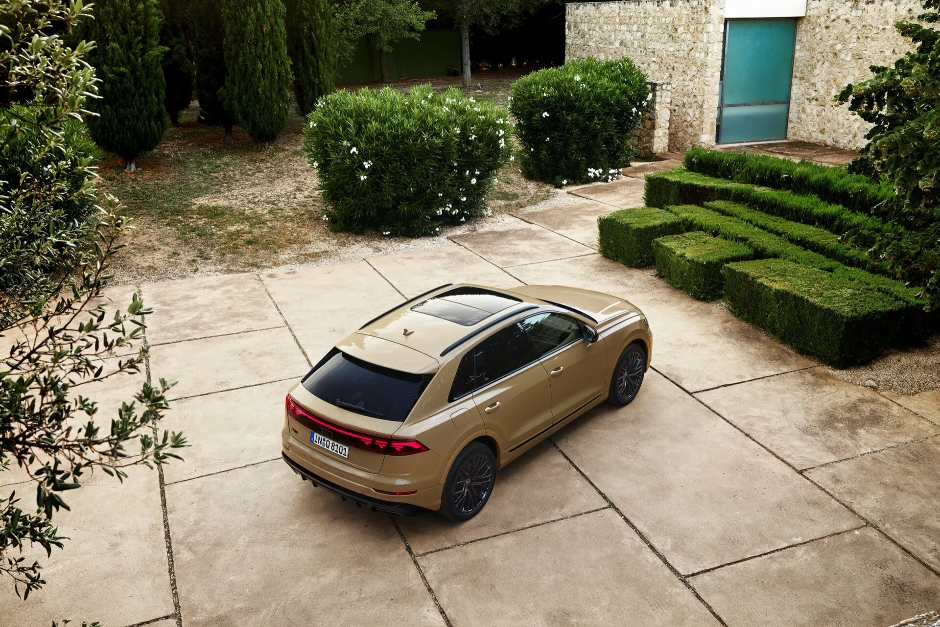 Sedan Audi Q8 2023 (4M-facelift) i hög vinkel, bakifrån och från sidan, visar LED-bakljus, panoramasoltak och sportiga fälgar, parkerad med en scenisk bakgrund av häckar och en stenbyggnad