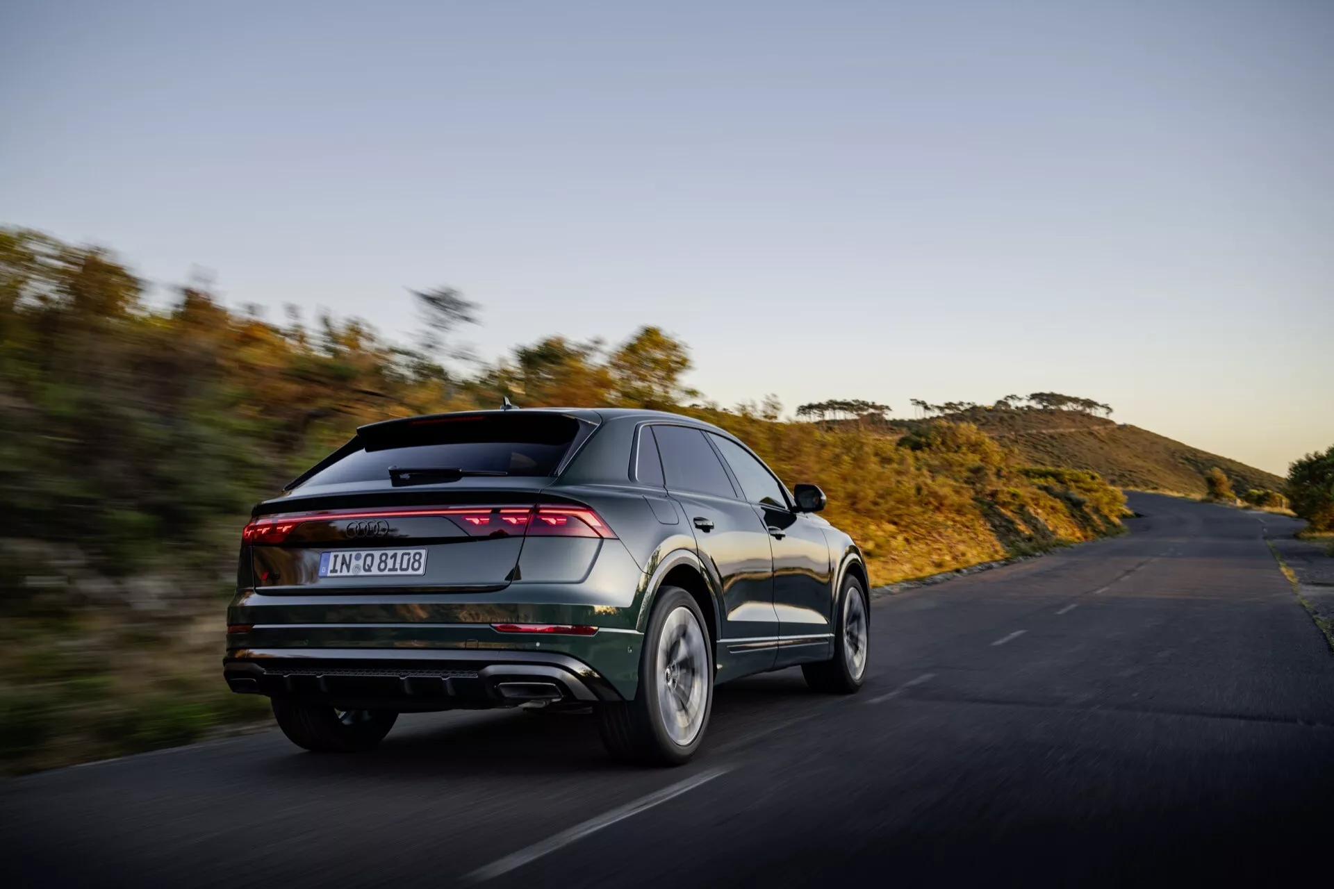 Audi Q8 2023 (4M-Facelift) i rörelse, fångad från en bakre trekvarts vinkel som visar fordonets moderna designfunktioner inklusive eleganta, fullbredds horisontella bakljus, dubbla avgasrör, bakre diffuser och integrerad spoiler mot en bakgrund av naturscen på en öppen väg vid solnedgång eller soluppgång, vilket belyser dess prestandaorientering och avancerad estetik.