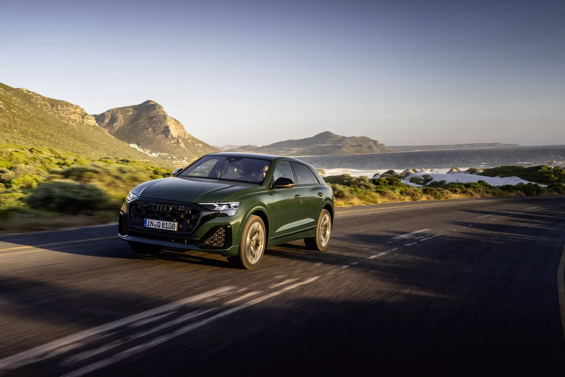 Audi Q8 4M-Facelift 2023 kör på en kustväg, markerad av sin aerodynamiska design, LED-strålkastare och tillverkarens emblem på grillen, fångad från ett främre trekvarts vinkel som visar lyx-SUV:en i rörelse mot en naturskön bakgrund