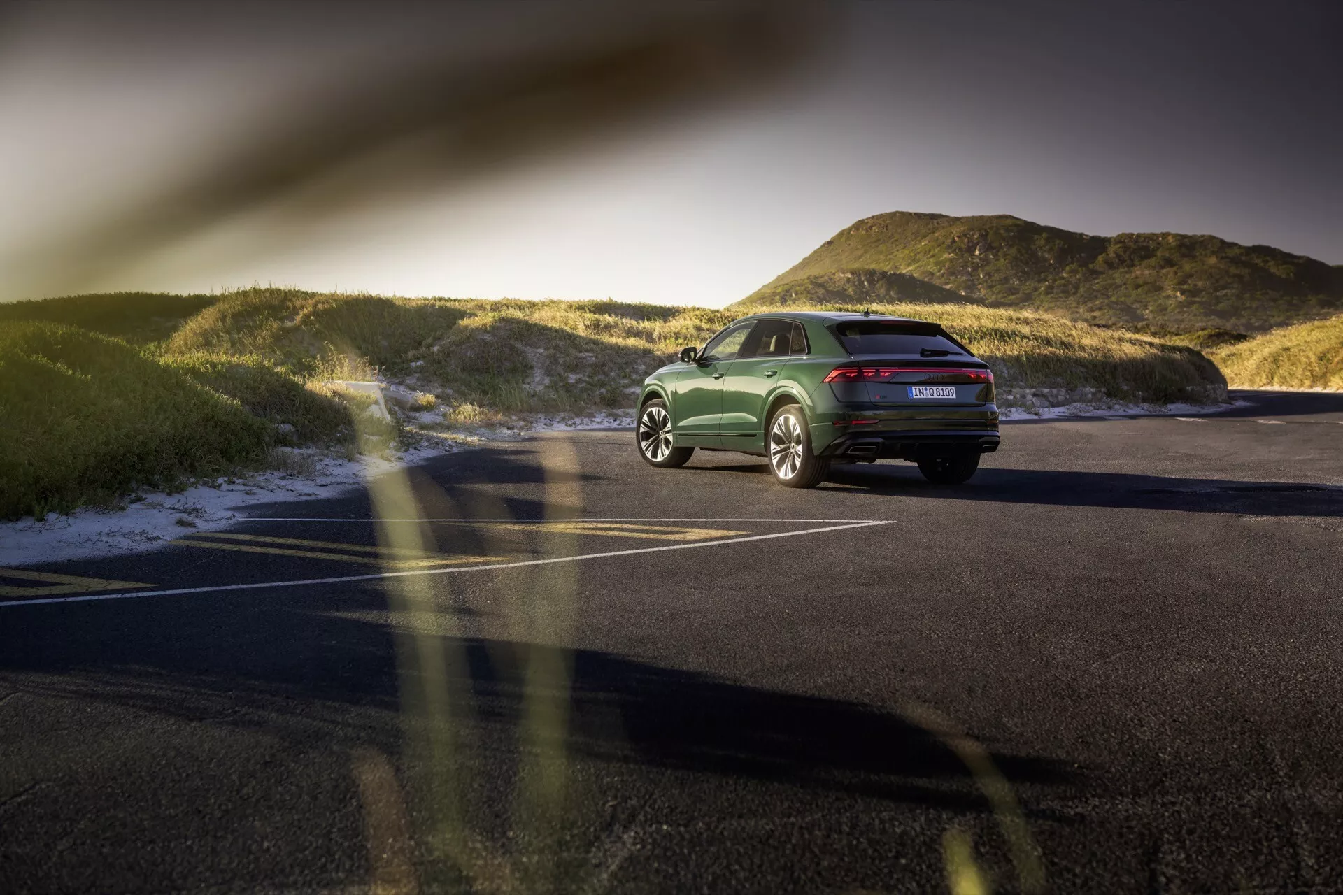 Bakre tredjedelssikten av Audi Q8 (4M-Facelift) 2023 på en asfalterad parkeringsplats vid solnedgången, som visar dess hållning, designlinjer, LED-bakljus, lättmetallfälgar och polerad grön finish med ett naturlandskap i bakgrunden.