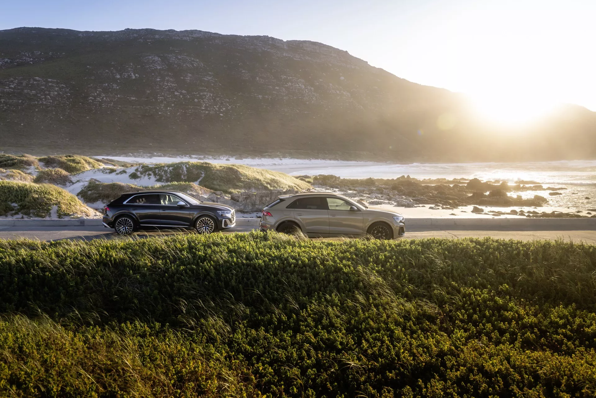 SUV och sedan Audi Q8 4M-Facelift 2023 parkerade under soluppgång eller solnedgång med hav och klippig strand i bakgrunden, med varmt sken och linsflare, visar deras aerodynamiska design, LED bakljus och reflekterande ytor