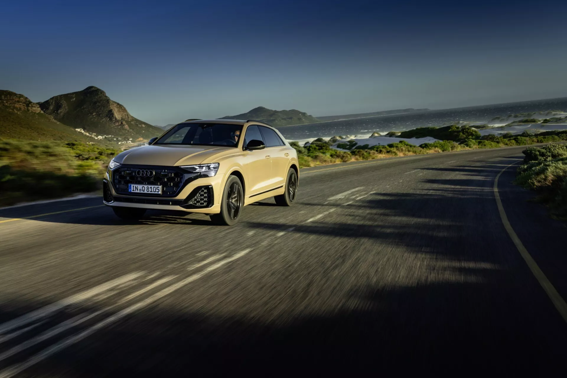 Audi Q8 4M facelift 2023 i rörelse på en kustväg, som framhäver den lyxiga bilens avancerade LED-strålkastare, framträdande grill, aerodynamiska motorhuv och dynamisk hantering, satt mot en bakgrund av berg och hav med fokus på design- och teknikfunktioner.