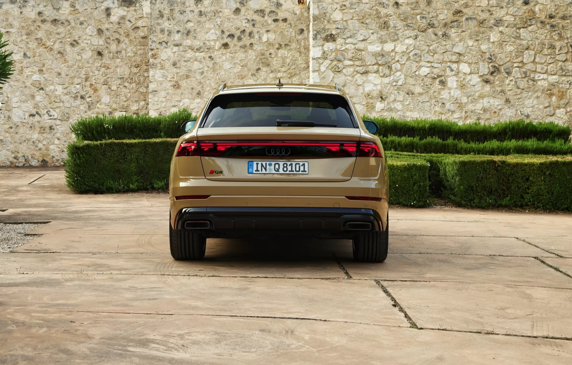 Audi Q8 2023 (4M-facelift) som visar bakifrån med moderna LED-bakljus, dubbla avgasrör, europeisk registreringsskylt, parkerad på ett stenlagt underlag mot en bakgrund av en stenmur och trimmade gröna häckar, vilket framhäver lyx och avancerade designfunktioner