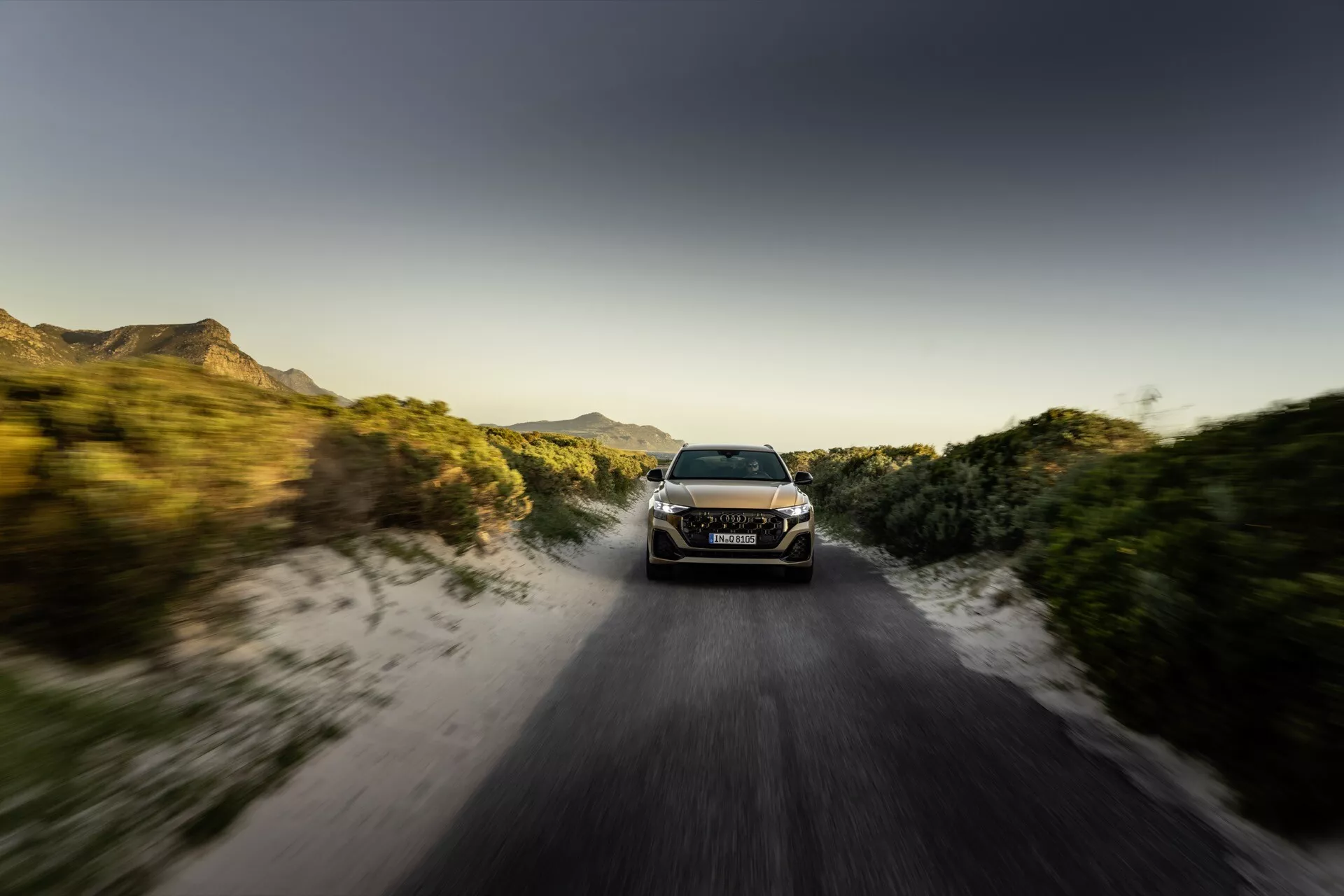 Audi Q8 2023 (4M-Facelift) i dynamisk rörelse på en oasfalterad väg vid soluppgång eller solnedgång med rörelseoskärpningseffekt, som visar sina LED-strålkastare, snygg aerodynamisk design och terrängkapacitet mitt i en lantlig miljö