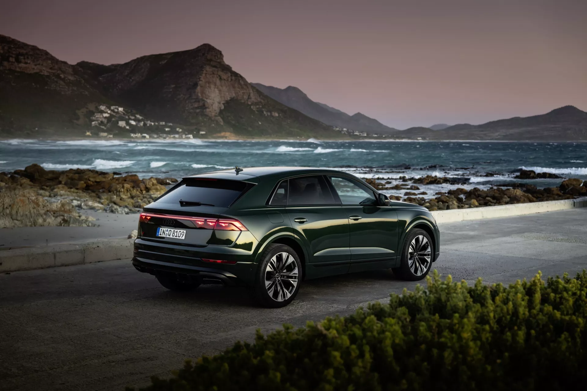 Audi Q8 2023 (4M-Facelift) parkerad vid en pittoresk kustplats vid gryning eller skymning som visar sin bakre kvartsutsikt, mörkt metalliskt grön finish och flerkantiga legeringshjul mot en lugn bergsbakgrund