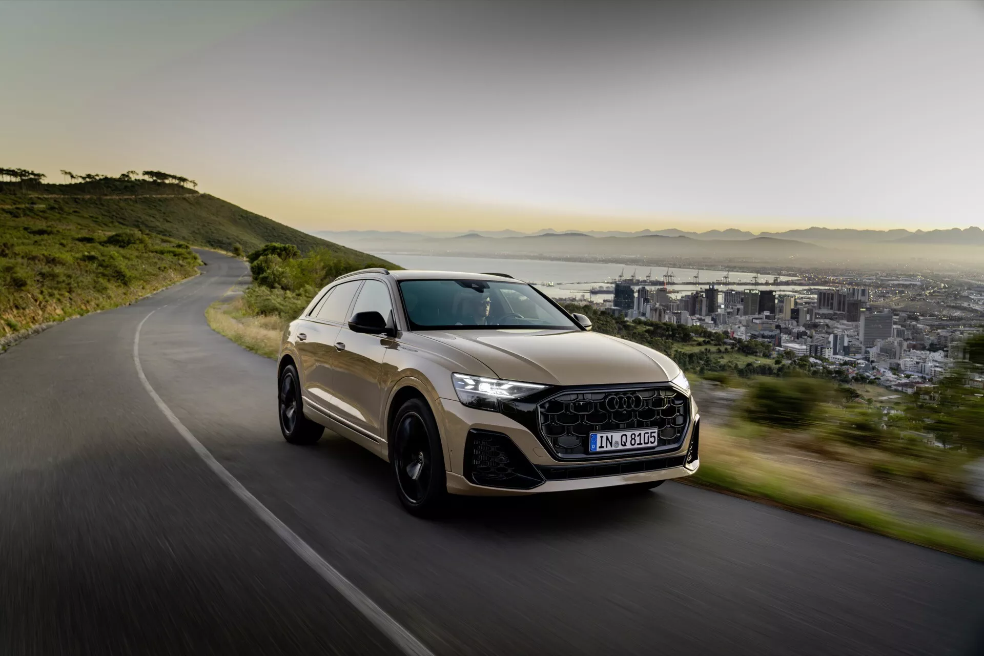 Lyxig crossover SUV Audi Q8 2023 (4M-Facelift) i rörelse på en slingrande väg, visar sportig design, LED-strålkastare, stora legeringshjul och ett panoramatak, mot en bakgrund av stad och berg i mjukt morgon- eller eftermiddagsljus.