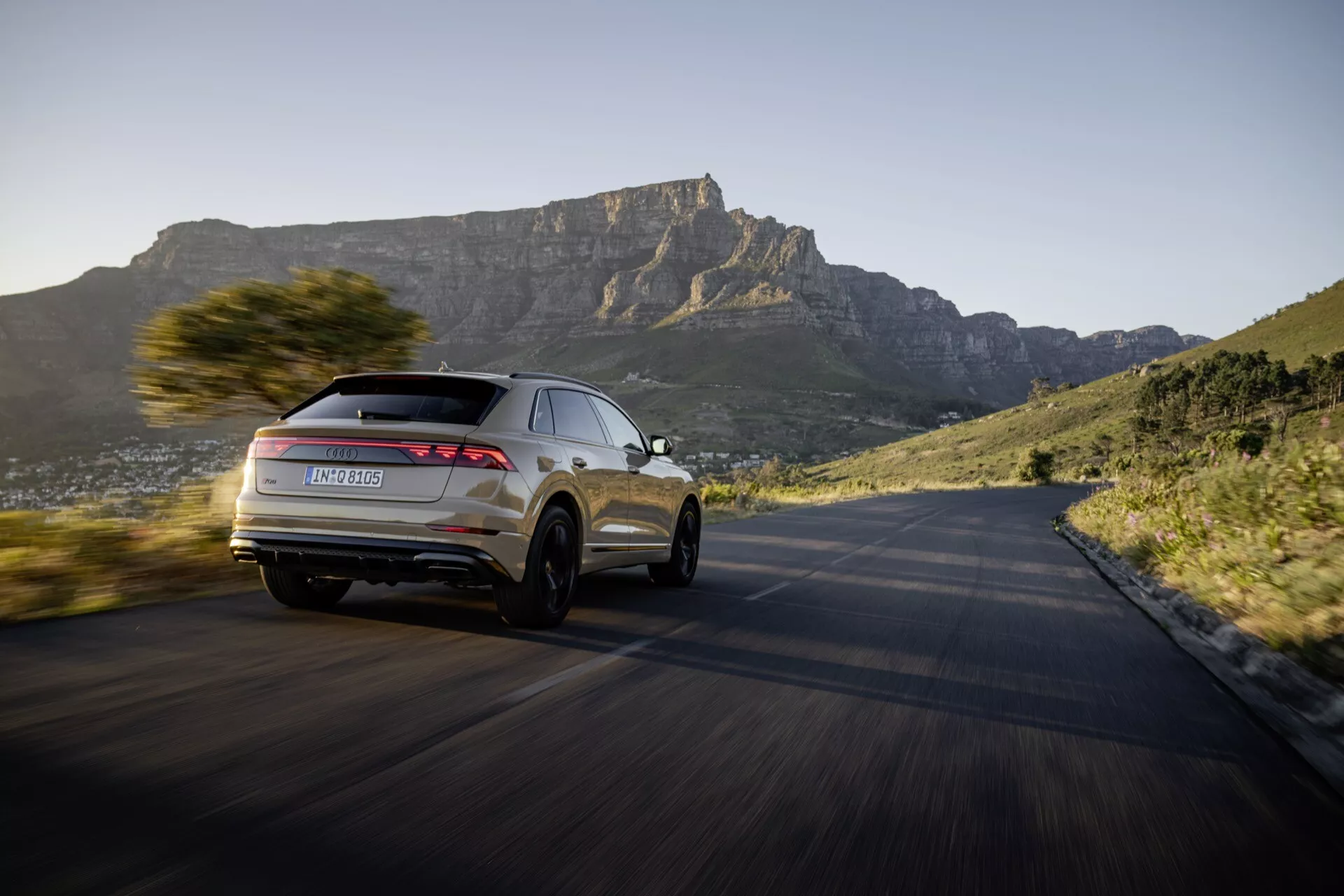2023 års Audi Q8 4M facelift-modell i dynamisk rörelse på en kurvig väg, framhäver dess moderna SUV-design och avancerade funktioner från ett bakre trefjärdedels perspektiv, med en naturskön bergig bakgrund vid solnedgången