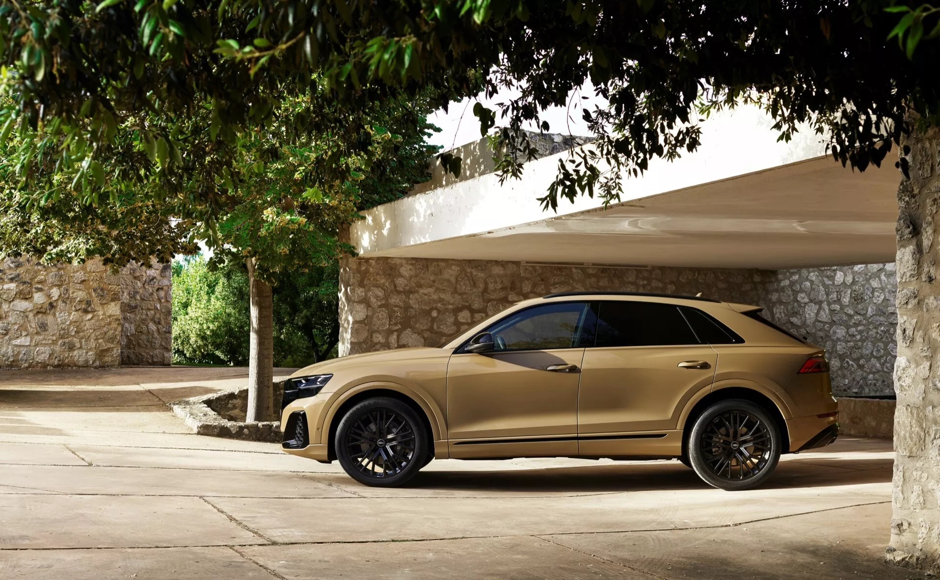 Audi Q8 2023 (4M-Facelift) i beige/guld, visar en fullständig sidovy med mörka legeringshjul, sidospeglar med integrerade blinkers, tonade fönster, infällda dörrhandtag och sluttande taklinje, parkerad i skugga på en solig dag