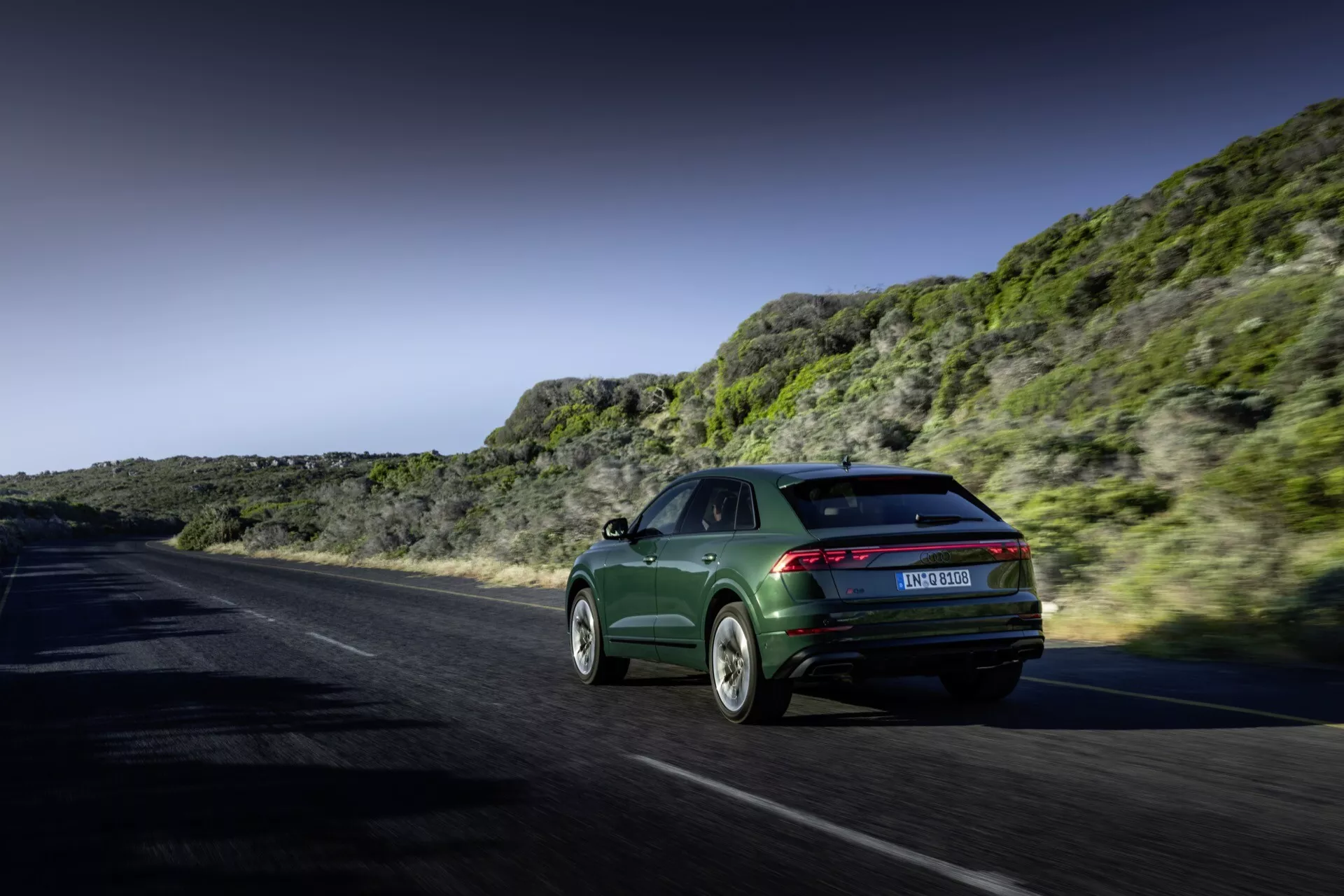 Audi Q8 2023 (4M-Facelift) i dynamisk rörelse på en kust- eller bergsväg, som visar sin sportback-stil, eleganta bakljus och sofistikerade legeringshjul mot en dramatisk landskap