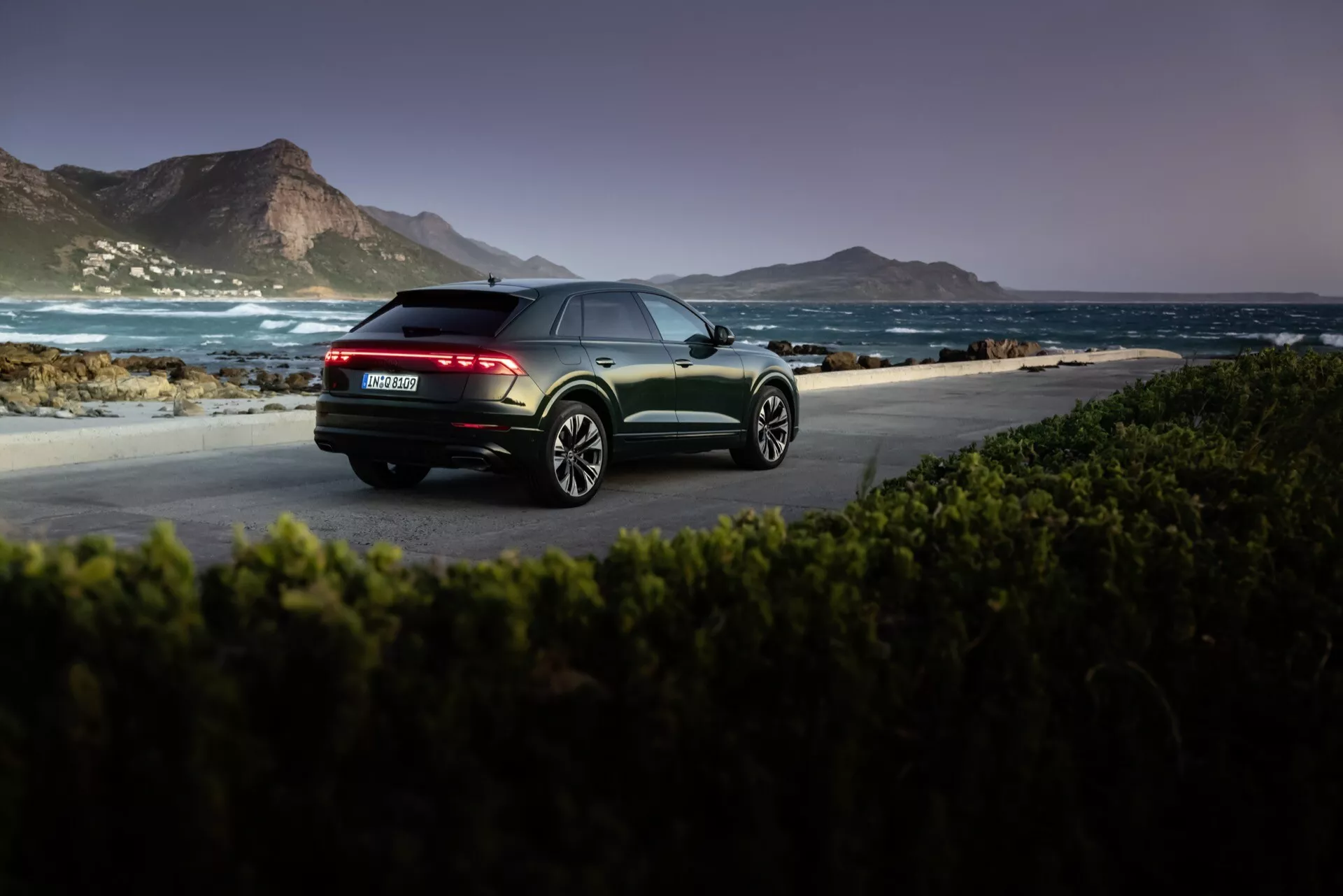 Audi Q8 4M-Facelift 2023 i metallfinish fångad vid gryning eller skymning i naturlig kustmiljö, framhäver vy från bakre trekvart med LED-bakljus, dubbla avgassystem och flerspakiga lättmetallhjul.