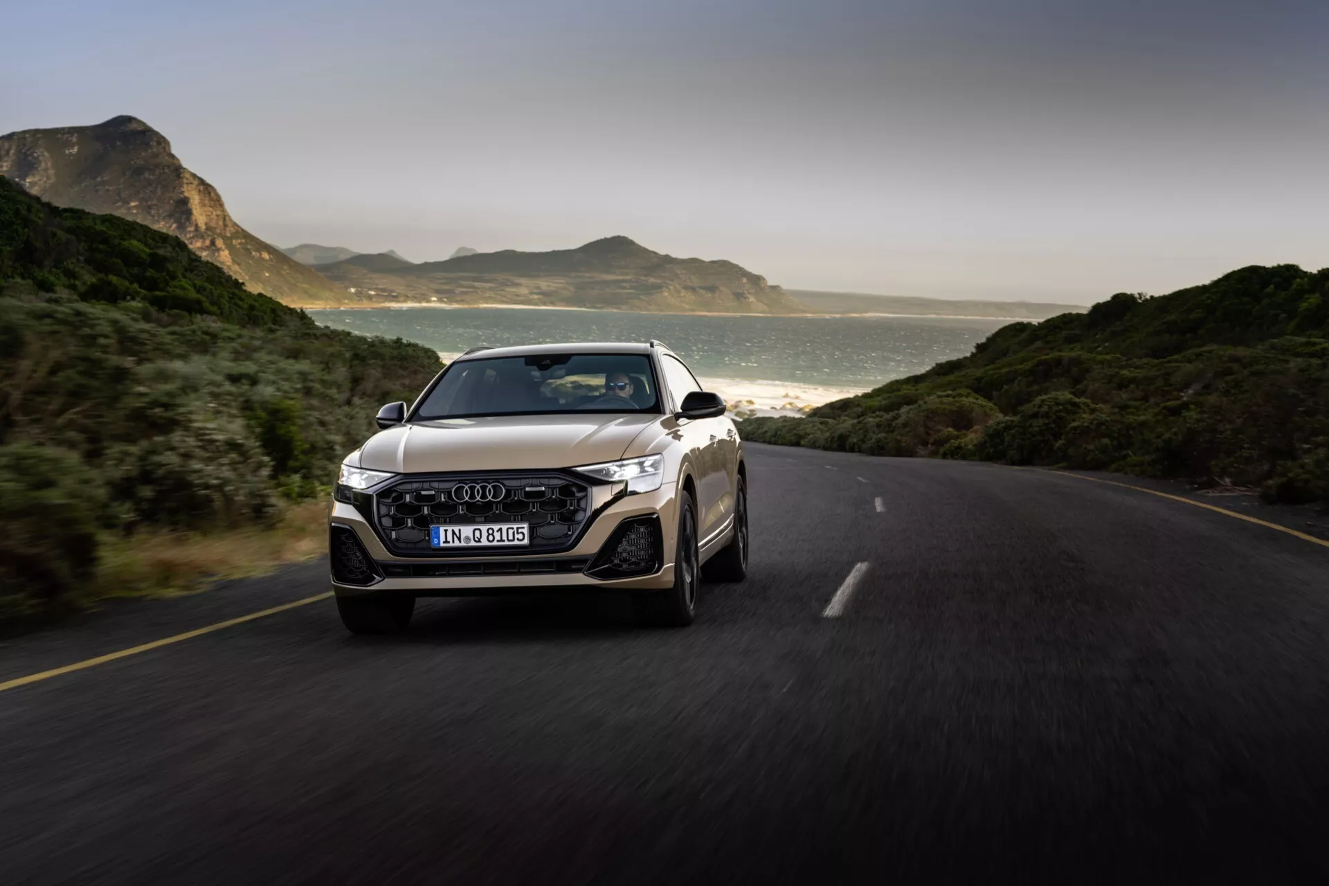 Audi Q8 2023 (4M-Facelift) fångad i rörelse på en asfalterad väg, som visar dess dynamiska design från en front tre fjärdedels vy framhävande grillen, LED-strålkastare, stötfångare och emblemet på grillen, med en naturskön bakgrund av berg och kustlinje i naturligt ljus