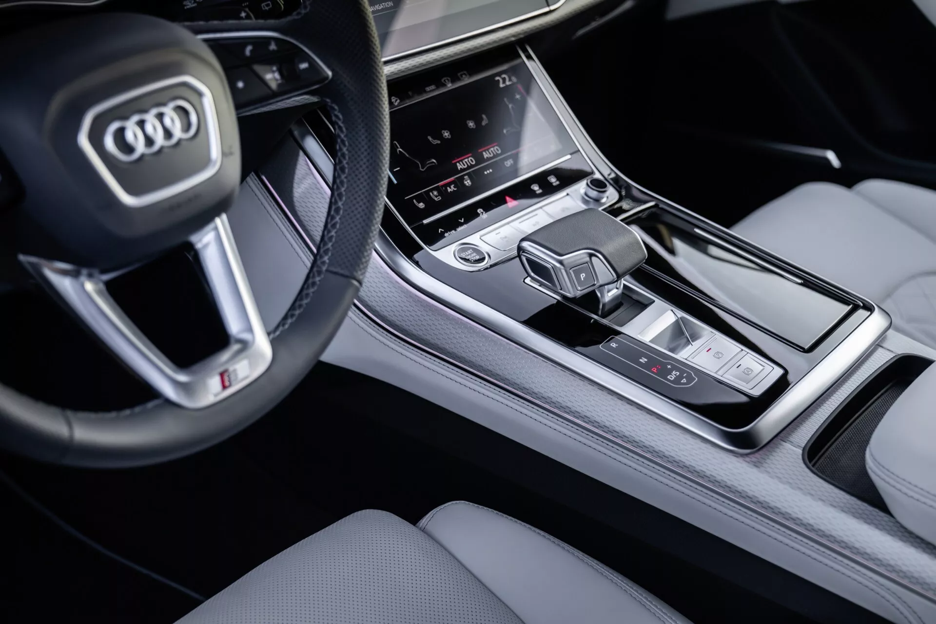 Närbild av mittenkonsolen och en del av ratten i en Audi Q8 2023 (4M-Facelift) från passagerarsidan, med fokus på knapparna på multifunktionsratten, den centrala infotainmentskärmen, kontrollområdet för luftkonditioneringen, och den moderna växelspaken inom en exklusiv interiörfinish.