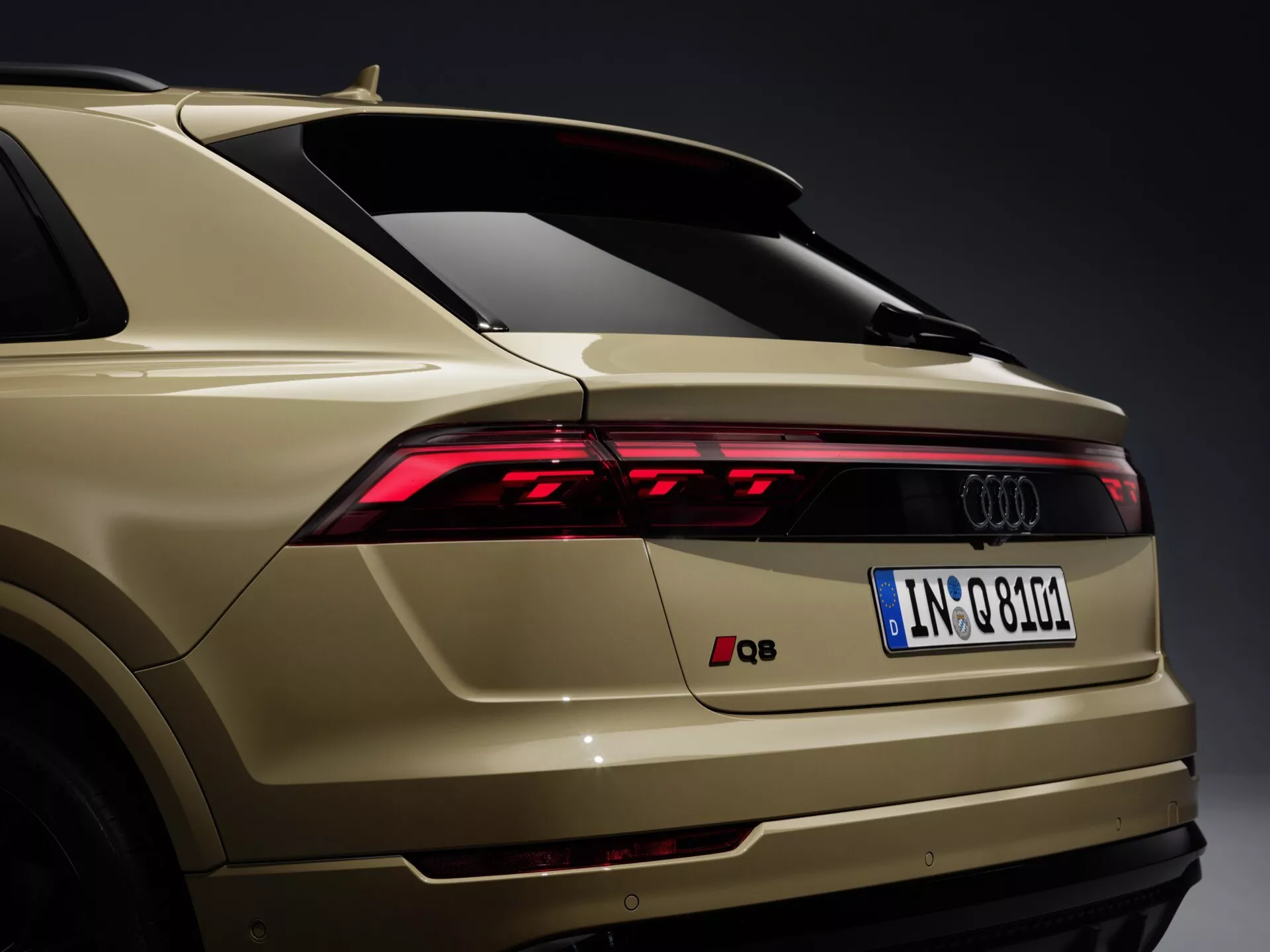 Bakre tre fjärdedels vy av en Audi Q8 4M-Facelift 2023 som visar LED-bakljus, snygg bakre stötfångare med integrerade sensorer, Audi-branding, europeisk registreringsskylt, bakspoiler, guld-beige lackfinish och hajfena antenn mot en neutral grå bakgrund.
