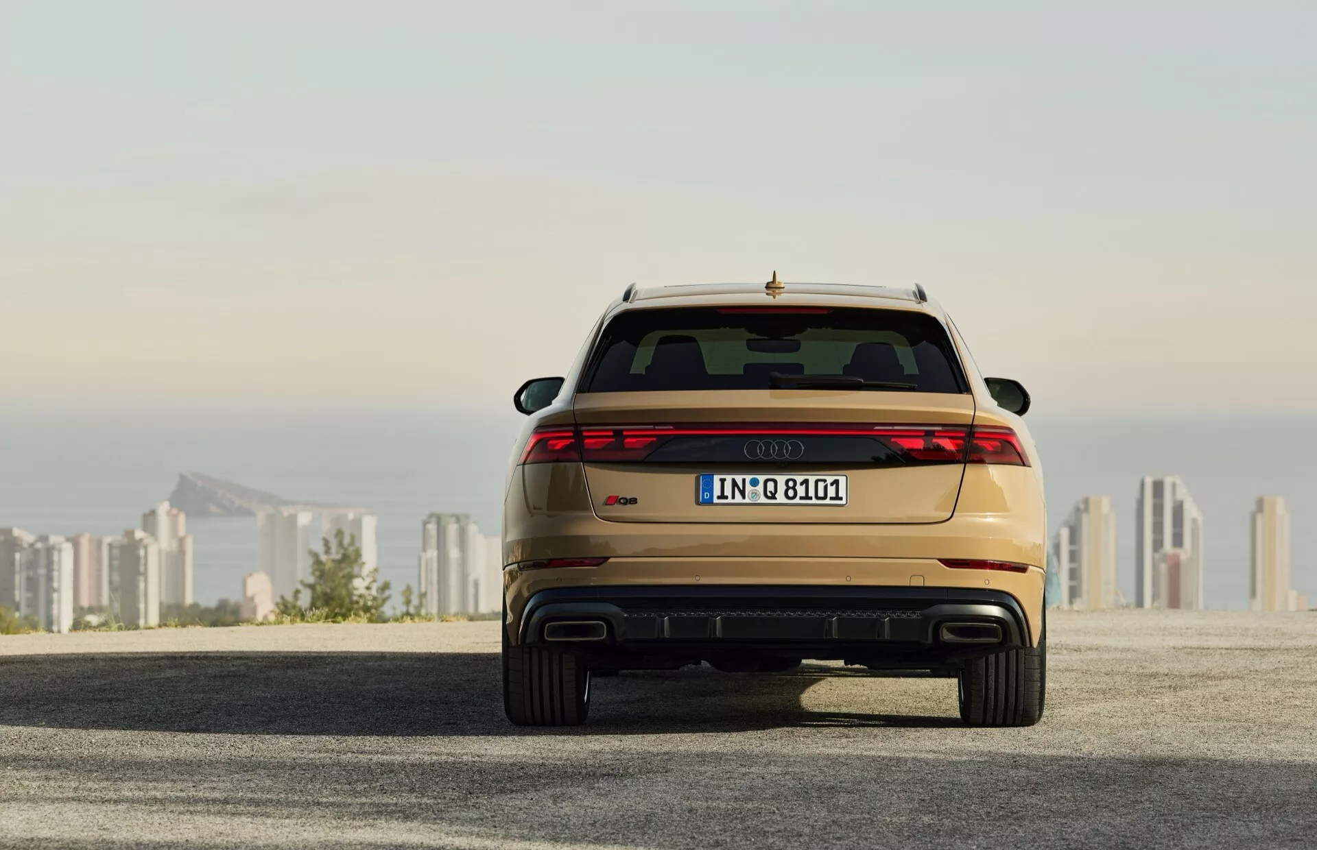 Baksida av Audi Q8 2023 (4M-Facelift) som visar LED-bakljus, dubbla avgasrör och metallicfärg, parkerad på en asfaltsyta med stadssilhuett i bakgrunden vid soluppgång/solnedgång