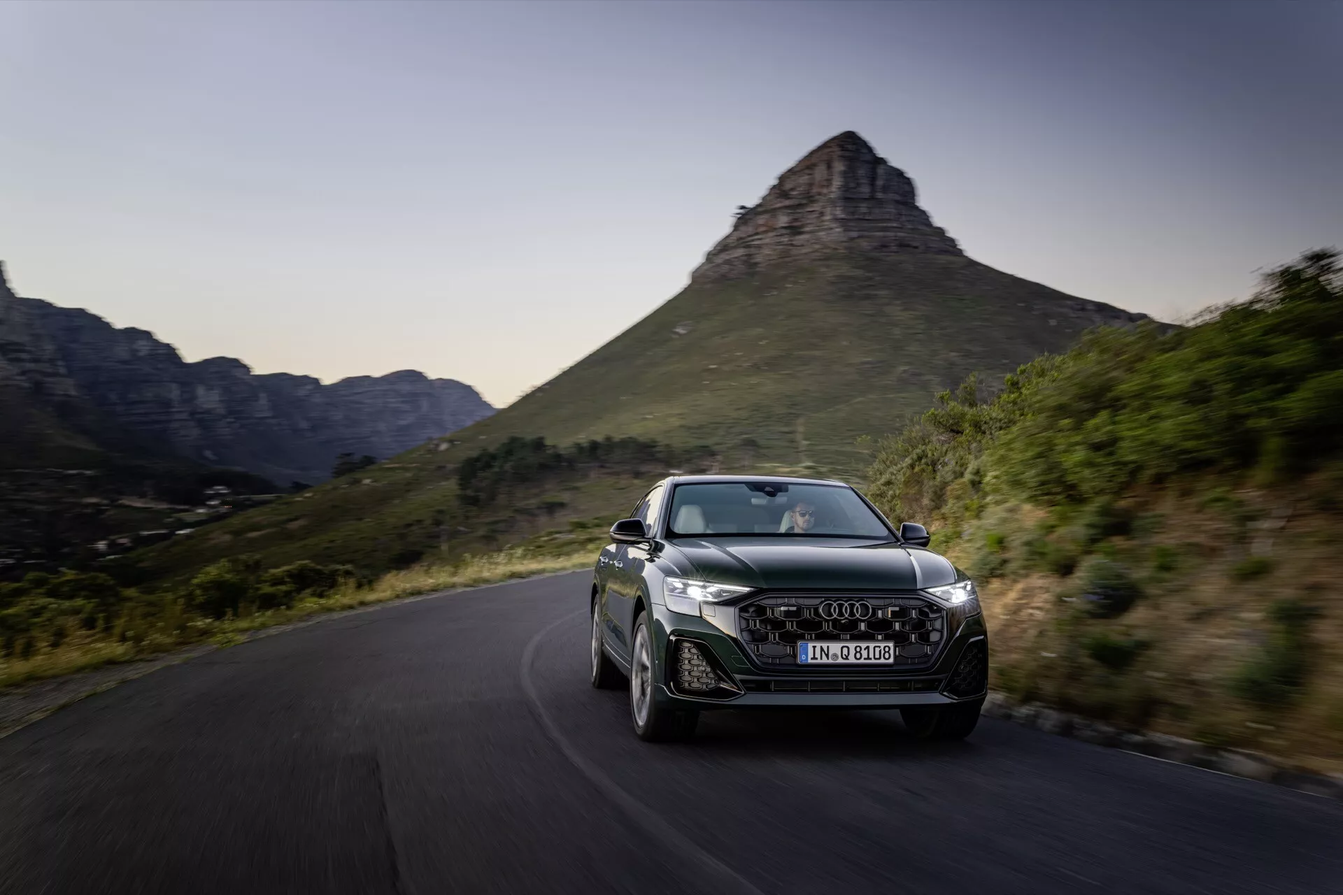 Audi Q8 2023 (4M-Facelift) i dynamisk rörelse på en naturskön slingrande väg, visar upp sin aerodynamiska design, LED-strålkastare, distinkta legeringshjul och integrerade luftintag mot en pittoresk bergsbakgrund under sen eftermiddag