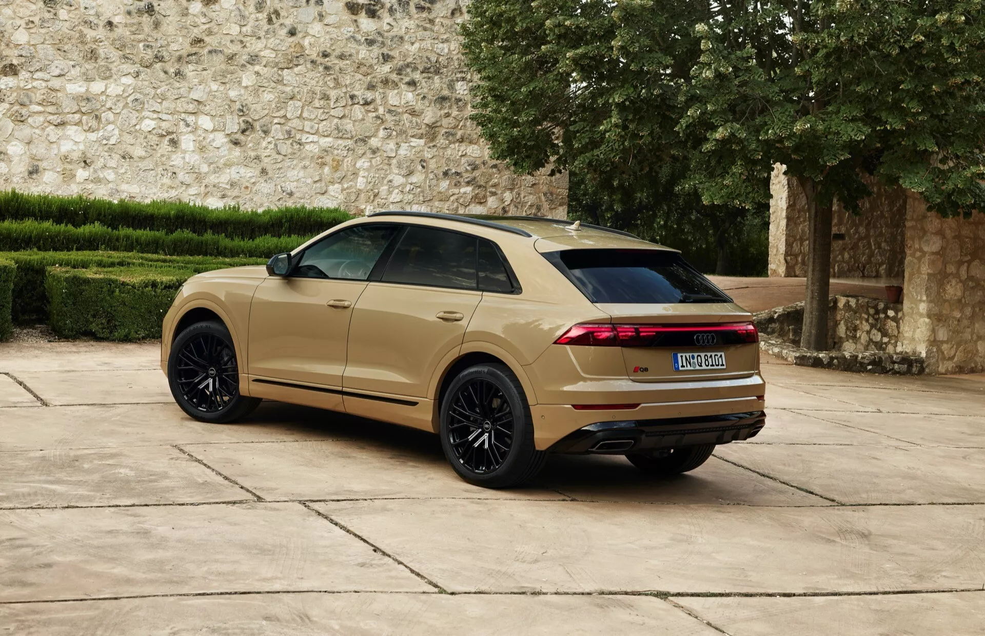2023 Audi Q8 (4M-Facelift) visas från ett tre kvarts bakperspektiv med stora svarta flerspakiga lättmetallhjul, metalliskt guldbegfärgad lack, förlängda horisontella LED-bakljus, framträdande bakspoiler och dubbla avgassystem mot historisk stenmur bakgrund