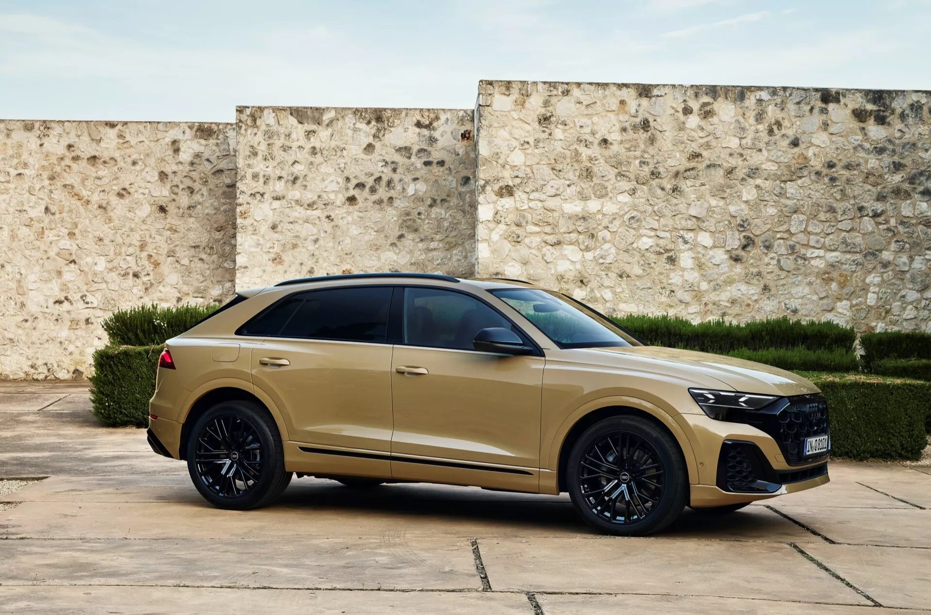 Trekvarts framifrån bild av en modern Audi Q8 2023 (4M-Facelift) i matt guld- eller beige-finish med en stor grill med bikake-mönster, LED-strålkastare, sportig frontstötfångare, svarta legeringshjul, lågprofildäck, infällda dörrhandtag, mörkt tonade fönster, en coupé-liknande sluttande taklinje, muskulös axellinje med vinklade bakljus och en takspoiler, placerad mot en texturerad vägg som tyder på en historisk eller samtida miljö.
