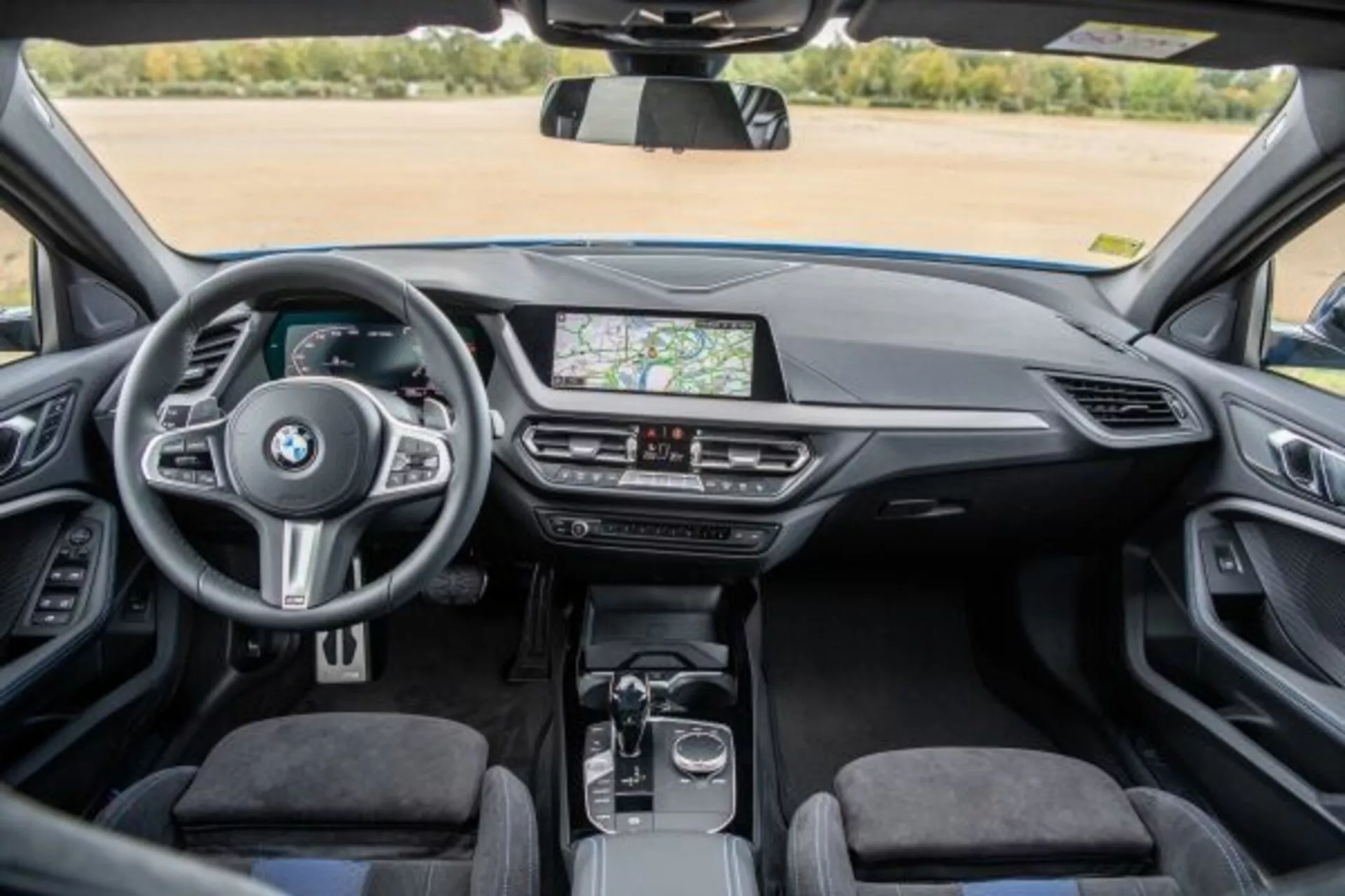 Interiörvy av BMW 1-serie M Performance 2019 från baksätets perspektiv som visar instrumentbrädan, mittkonsolen och ratten med monterade kontroller, pekskärms infotainmentsystem som visar GPS-navigation, luftventiler, klimatkontroller, automatisk växelspak, roterande ratt och pekplatta på mittkonsolen, premiumklädsel och en klar, välbelyst kupé