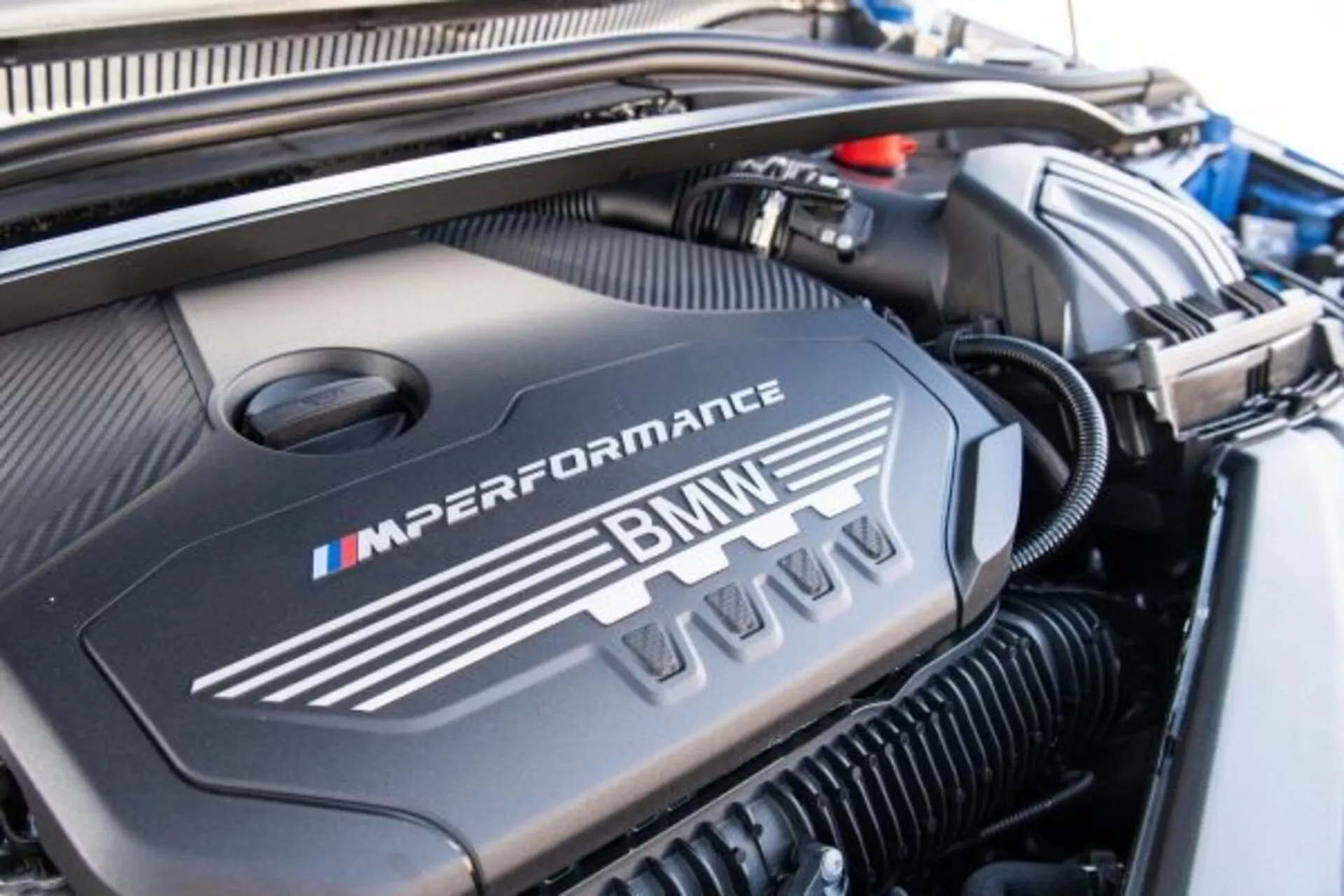 Närbild av motorutrymmet i en BMW 1-serie M Performance från 2019 med motorkåpan märkt med M-logotypen i dess signaturfärger blått, lila och rött, vilket framhäver den sportiga, högpresterande designen.