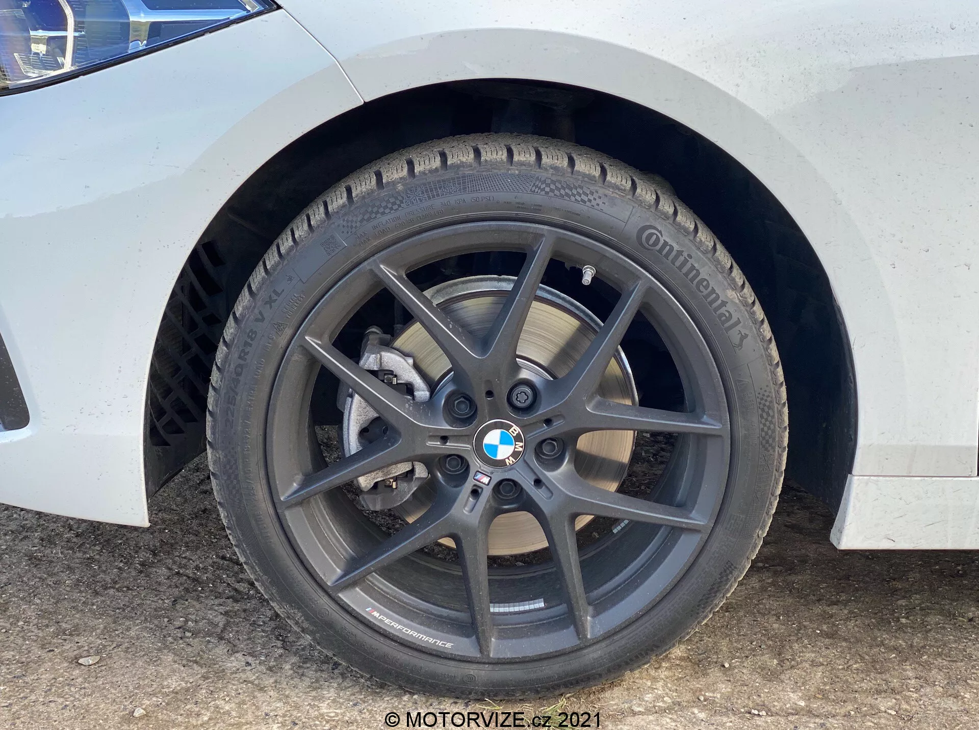 Närbild på framhjulet och framskärmen på en BMW 1-serien 2019 (BMW-116i). Perspektivet på marknivå framhäver den mörkgrå femekrade lättmetallfälgen med BMW-emblem, däck av märket Continental med synligt mönster, ventilerad bromsskiva och silverfärgad enkolvs bromsok. Den vita lacken med smuts- och vattenfläckar tyder på användning.