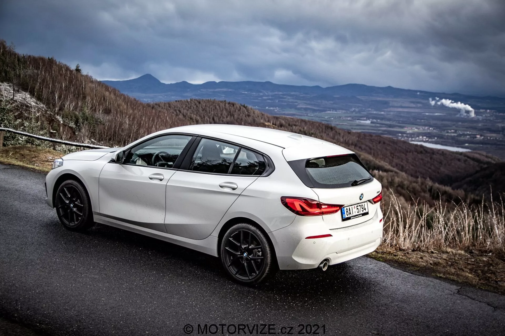 En vinklad bakre sidovy av en vit 2019 BMW 116i hatchback på en asfalterad väg med en dramatisk molnig himmel. Bilden framhäver fordonets bakre design, bakhjulsbågar, mörkfärgade lättmetallfälgar, dubbelt avgasrörsystem och sidoprofil inklusive bakre passagerardörr och förardörr. Miljön antyder en kullig eller bergsväg och betonar bilens sportiga och dynamiska attribut. Fotografens vattenstämpel 'MOTORWIZE.cz 2021' är närvarande.