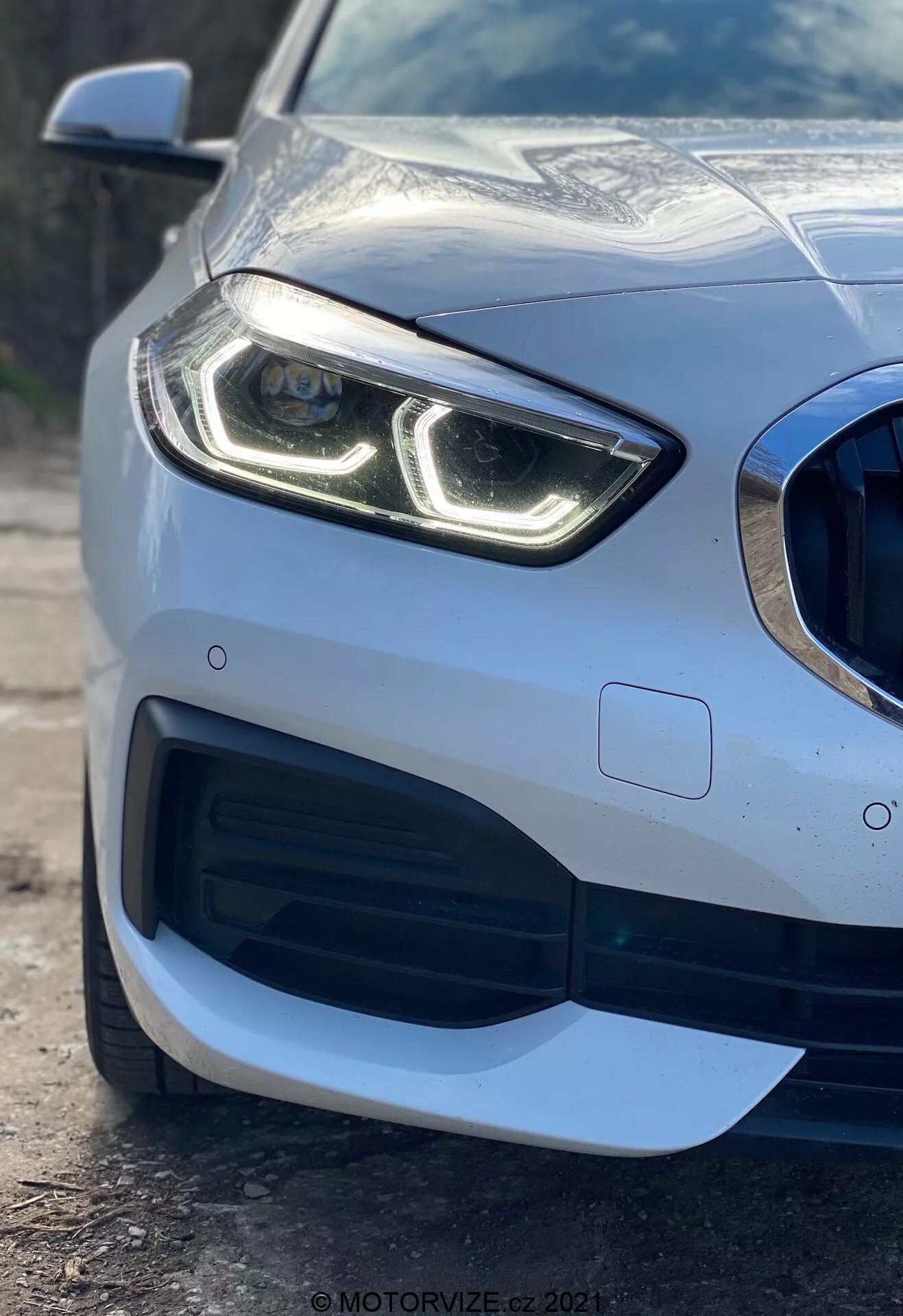 Närbild av den vänstra främre delen av en BMW 1-serie 2019 (BMW 116i) som framhäver dess moderna strålkastare med distinkta LED-dagljuslampor, grill i bikupemönster med stor luftintag, sofistikerad aerodynamisk design på främre stötfångaren och parkeringssensor inbyggd i stötfångaren, vilket visar den premium metalliska lacken och reflektiva karossen.