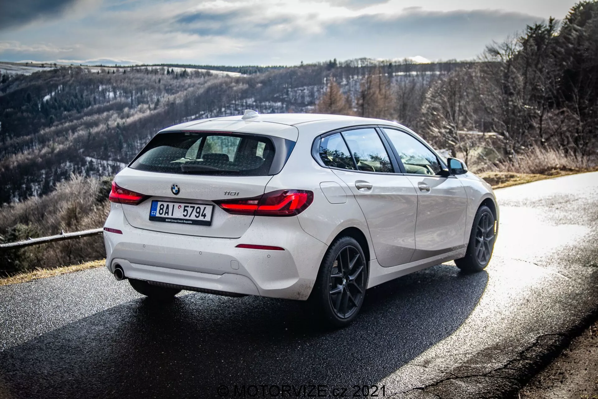 En dynamisk trekvart bakifrån vy av en vit BMW 116i 2019, som visar dess högra sida och bakre del under dagsljus med tydliga solstrålar och skuggor. Bilden betonar designdetaljer, inklusive svarta accenter på bakre diffusorn, hjulfälgar och sido spegelkåpor, kompletterad med legeringshjul och ett dubbelt avgas system, vilket antyder en sportigare variant. Vägen svänger åt höger med en backig eller bergig bakgrund, idealisk för körentusiaster.