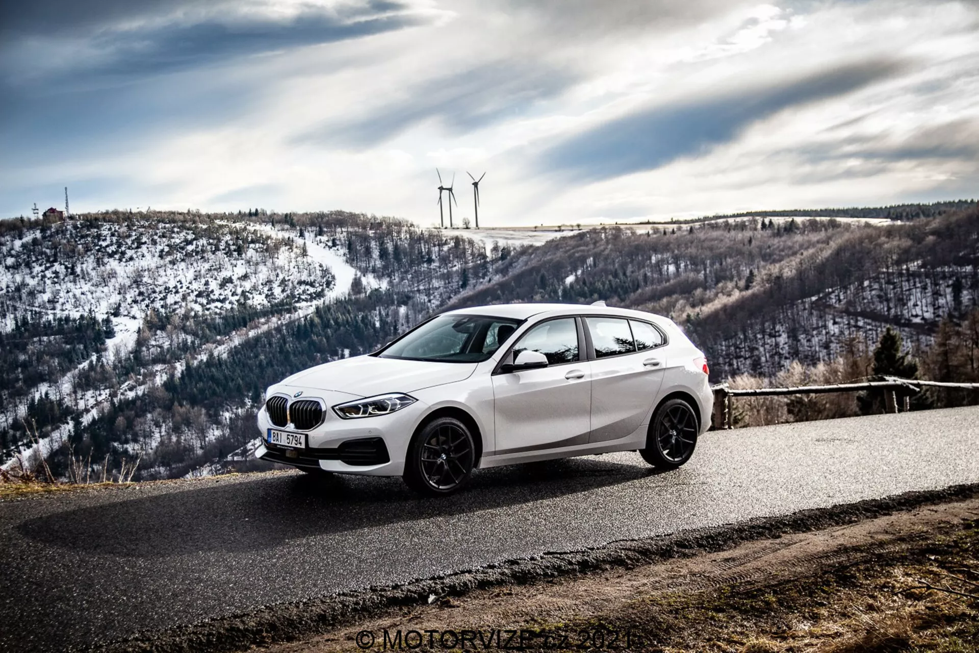 En vit BMW hatchback, troligen en BMW 1er-Reihe 2019 (BMW 116i), är parkerad på en kurvig asfaltsväg omgiven av ett kuperat, snötäckt landskap med vindkraftverk. Fotot, taget från en sido-frontvinkel, framhäver bilens frontgrill, strålkastare, aerodynamiska linjer och lättmetallfälgar mot en mulen himmel.