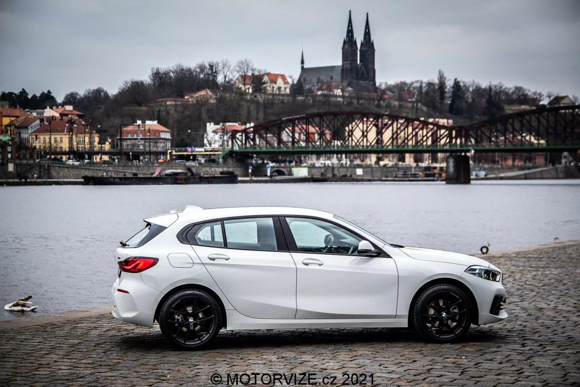 En vit BMW 1-serie hatchback från 2019 (BMW-116i) parkerad på en kullerstensyta nära en vattenkropp med ankor, visande en sidoprofil med lätt bakre kvadratisk vy. Bilen har dynamiska skulpterade karaktärslinjer, sluttande taklinje, svarta flerekrade lättmetallfälgar och synliga strålkastare samt sidospeglar. I bakgrunden finns en bro, byggnader på en kulle och arkitektoniska element som antyder en europeisk miljö. Belysningen är diffus under mulen himmel och bilden innehåller en vattenstämpel © MOTORVIZE.cz 2021.