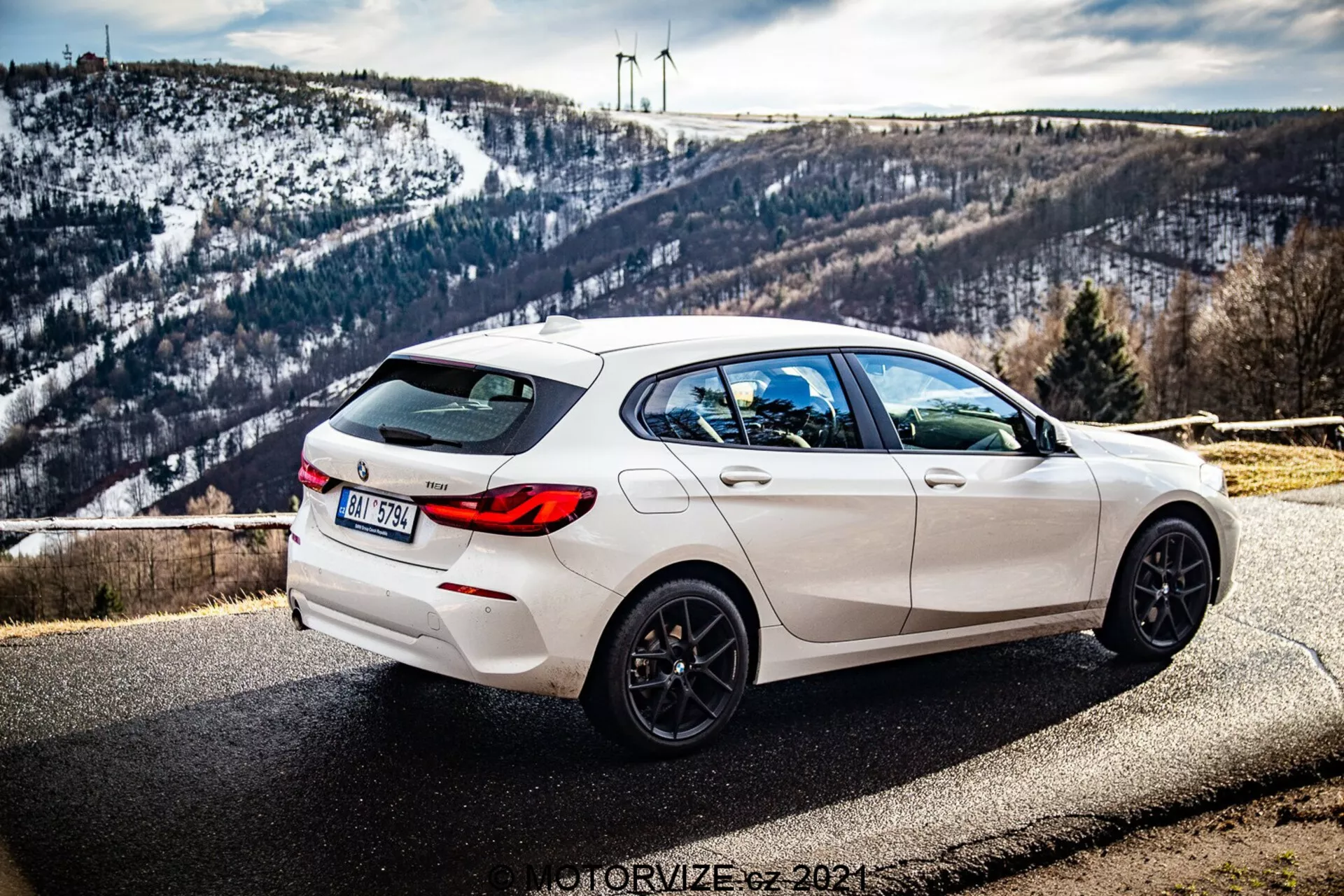 Bilden visar en vit BMW 1-serie (BMW-116i) 2019-modell parkerad vid vägrenen med en naturskön bakgrund som inkluderar ett kuperat, snötäckt landskap och vindkraftverk i horisonten. Fotot är taget från en trekvarts bakvinkel, vilket tydligt visar bilens högra sida. Utsikten framhäver de eleganta designlinjerna, den bakre spoilern, de femekrade svarta lättmetallfälgarna, dubbla avgasutsläpp och mörka tonade rutor. Bilen har även en bakre vindrutetorkare och en integrerad hajfensantenn på taket. Skärpedjupet behåller bilen i skarpt fokus med dagsljus som framhäver dess sportiga estetik medan bakgrunden förblir subtilt detaljerad.
