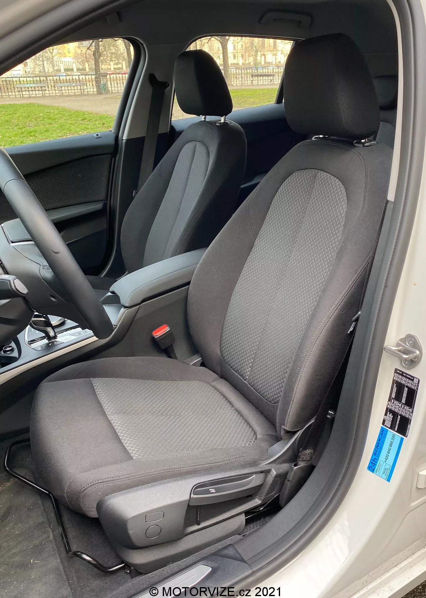 Närbild av interiören i BMW 1er-Reihe 2019 (BMW-116i) med fokus på förarsätet med en modern tvåtonstygdesign, huvudsakligen svart med ljusare grå mönster, tagen från en sned vinkel med bildörren helt öppen. Sätet har en nackstöd mot ljuset, vilket skapar en silhuetteffekt runt kanterna, och inkluderar en spak för manuella justeringar framåt eller bakåt, samt en liten svart knapp nära kanten av bilens interiör. Det naturliga dagsljuset framhäver den skarpa fokuseringen på sätet, med en suddig bakgrund som visar ratten och instrumentbrädan.