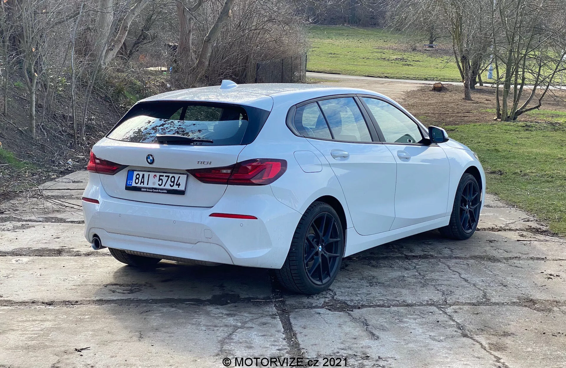 Bakre trekvarts vy av en vit BMW 1-serie kombikupé (2019 års BMW 116i-modell) parkerad utomhus i dagsljus. Bilens sidoprofil och baksida är tydligt synliga, med BMW-emblem, modellbeteckning, distinkta wrap-around bakljus, en bakspoiler, svarta fälgar, kombikupé karossstil, dubbla avgasrör, bakre vindrutetorkare, lätt tonade fönster och backspeglar med integrerade blinkers. Bilen är placerad på ett betongområde med gräsplättar och en landsbygd- eller förortsinställning i bakgrunden.