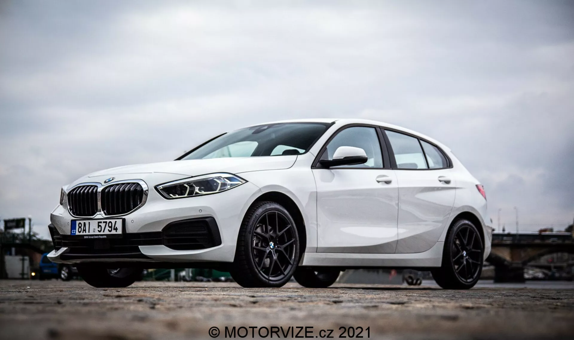 En vinklad sidovy av en vit BMW 1-serie 2019 (BMW 116i), med framträdande designelement som en njurformad grill, eleganta LED-strålkastare och en främre stötfångare med luftintag och möjliga radarsensorer för förarassistanssystem. Fotot fokuserar på lättmetallhjulen och den nedre delen av fordonet, vilket framhäver den sportiga flerradiga svarta lättmetalldesignen och den högeffektiva skivbromsen. Hatchbackens karaktärslinjer och aerodynamiska skulptering betonar ytterligare dess moderna och dynamiska exteriör.