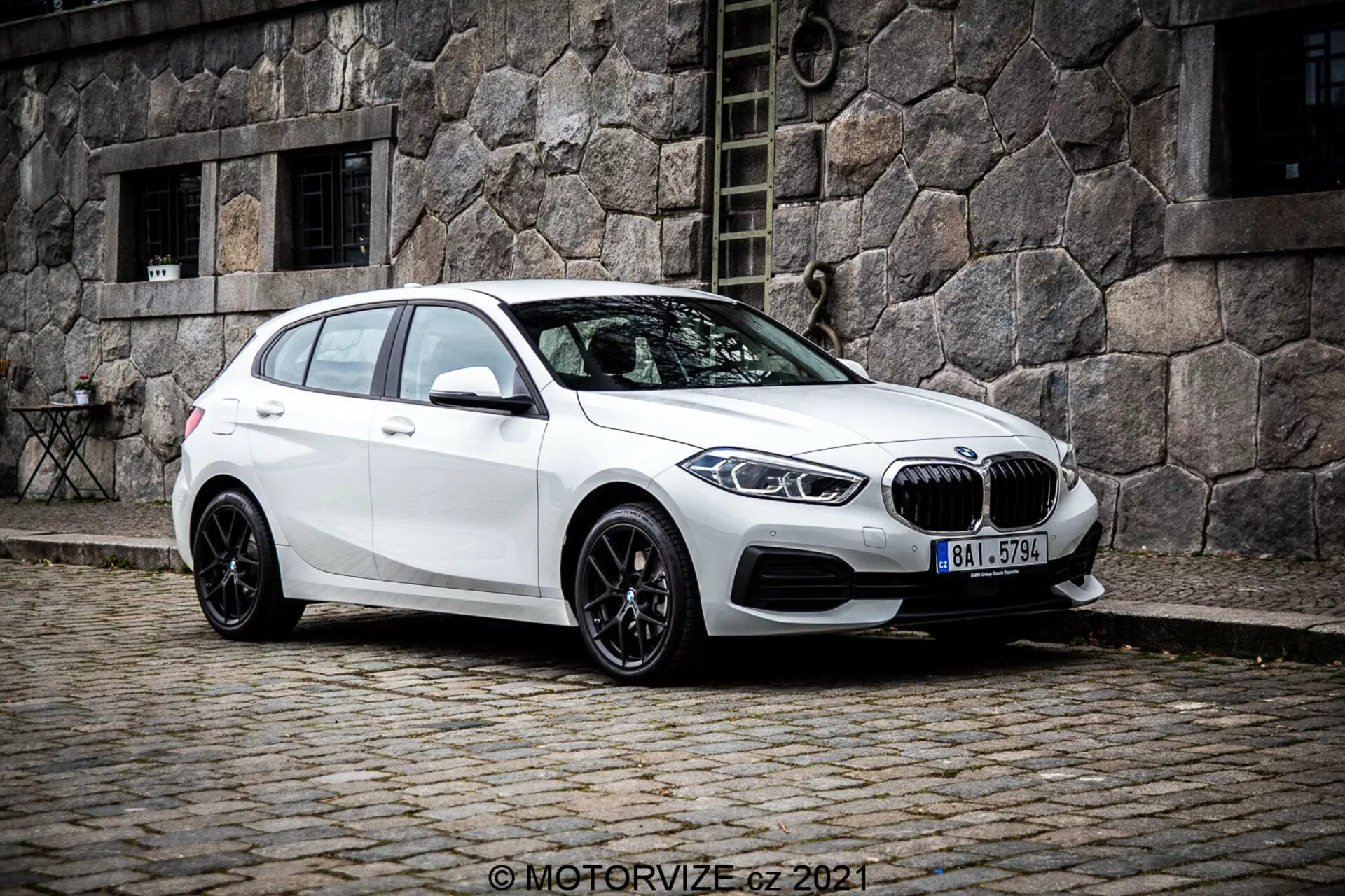 En vit BMW 1er-Reihe 2019 (BMW 116i) halvkombi sedd från en trekvarts frontal vinkel, som visar vänstra sidan av fordonet. Bilden, tagen från en något lägre vinkel, framhäver bilens hållning och designlinjer. Den är placerad på en kullerstensgata med en gammal stenbyggnad i bakgrunden, vilket skapar en klassisk urban scen. Den karaktäristiska njurgrillen och BMW-emblemet är tydligt synliga på fronten, omgiven av eleganta, vinklade LED-strålkastare. Bilen har mångekrade svarta lättmetallfälgar, en sluttande taklinje, sidospeglar i karossfärg med integrerade blinkers och tonade fönster för bakre passagerare.