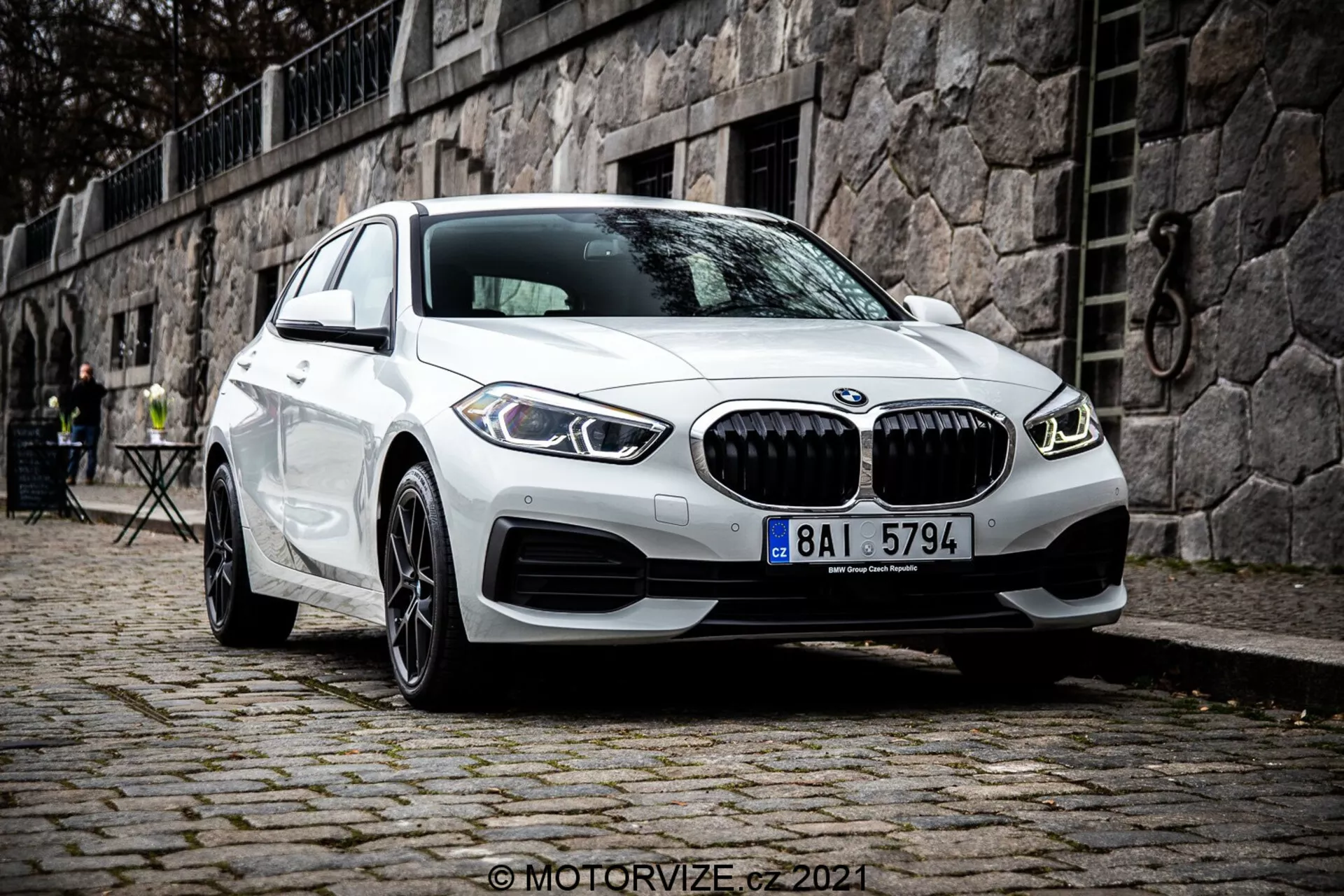 Modern vit BMW 1-serie (BMW 116i) 2019 årsmodell med den karakteristiska njurgrillen, dubbla cirkulära LED-varselljus och sportig främre stötfångardesign, parkerad på kullerstensyta med ett historiskt europeiskt landskap i bakgrunden som innehåller en stenmur och utomhuscaféets möbler.