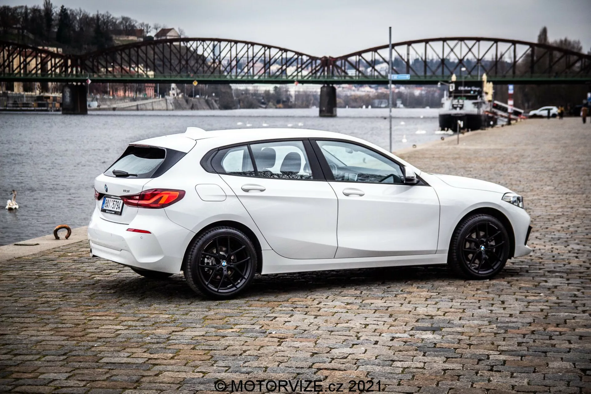 Bild av en vit 2019 BMW 1er-Reihe 116i kombi från en trekvarts bakre vy, som betonar dess eleganta, moderna design med sportig hållning, svarta flerspoksfälgar, lätt tonade fönster och praktiska funktioner som bakrutedefroster och torkare. Bilen visar skarpa linjer och konturer, dubbla avgasrör, subtil bakre takspoiler, backkamera och parkeringssensorer. Den är parkerad på en kullerstensyta nära en vattenkropp med en urban bro i bakgrunden, vilket antyder en livsstil i utomhusmiljö.
