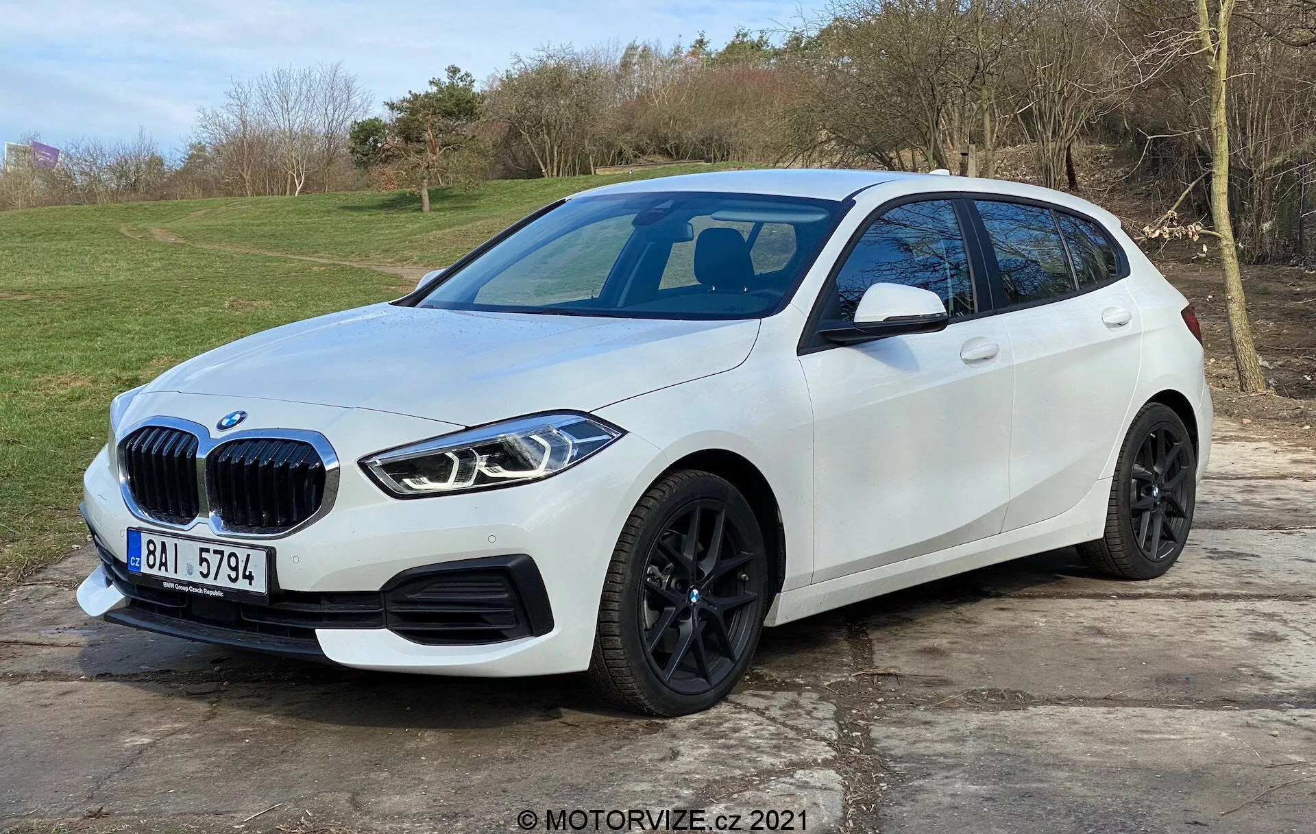 Bilden visar en modern vit BMW 1-serie (BMW 116i) hatchback placerad utomhus med en naturlig, gräsbeklädd bakgrund. Fotograferad från en trekvartsvy framifrån som favoriserar förarsidan, framhäver den fordonets karakteristiska njurgrill, eleganta kantiga LED-strålkastare, svarta lättmetallhjul, väldefinierad sidskarv och integrerade blinkers på sidospeglarna. Främre stötfångaren har parkeringssensorer medan ett panoramaglastak och eventuell framåtvänd kamera kan antydas. Bilen är parkerad på ett stenlagt område och upptar största delen av ramen, vilket antyder modern bildesign och avancerade teknikfunktioner.