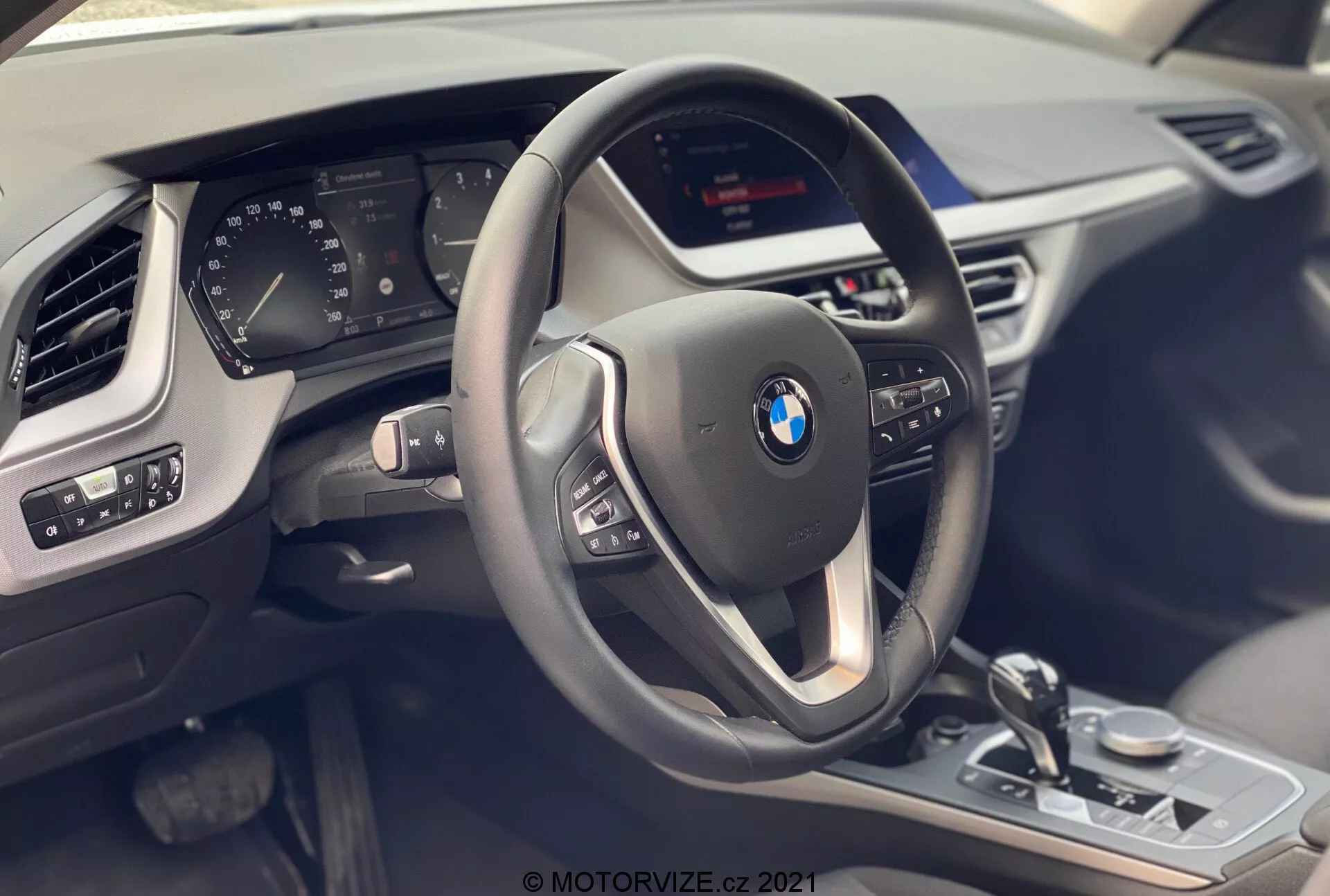 Interiörvy av BMW 1-serien 2019 (BMW 116i) från passagerarsätet, som framhäver förarens cockpit med en modern tre-ekrad ratt med multifunktionsknappar och läderöverdrag med kontrasterande sömdetaljer. Bakom ratten är en delvis synlig digital instrumentpanel som visar olika fordonets parametrar. Mittkonsolen rymmer en osynlig infotainmentskärm och synliga kontroller som växelväljaren, iDrive-kontrollreglaget, körlägesväljaren och parkeringssensorernas kontroller, alla med metalliska accenter och en blank svart finish. På förarsidans dörrpanel visas spegeljusteringar och fönsterkontroller. En del av passagerarsidans luftventilationsgaller och texturen på instrumentpanelen är också synliga, vilket betonar lyx, teknik och modern design.