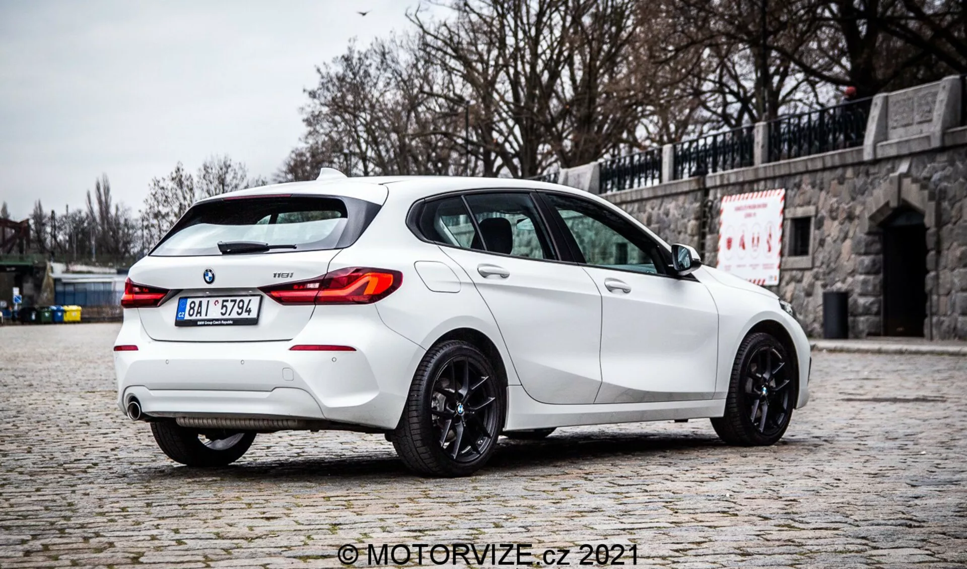 En högkvalitativ fotografi av en vit BMW 1-serie 2019 128ti hatchback tagen ur ett bakre trekvartsperspektiv, som betonar höger sida. Bilen står parkerad på en kullerstensbelagd gata med en stenmur i bakgrunden, vilket framhäver dess sportiga design med en bakre takspoiler, dubbla avgasrör, bakre diffusor, LED-bakljus, tonade rutor och mörkfärgade lättmetallfälgar. Den diffuserade belysningen, avsaknaden av hårda skuggor och de tydliga detaljerna antyder en molnig miljö, vilket ger en autentisk återgivning av fordonets färg och funktioner. Bilden har vattenstämpeln 'MotorVize.cz 2021'.