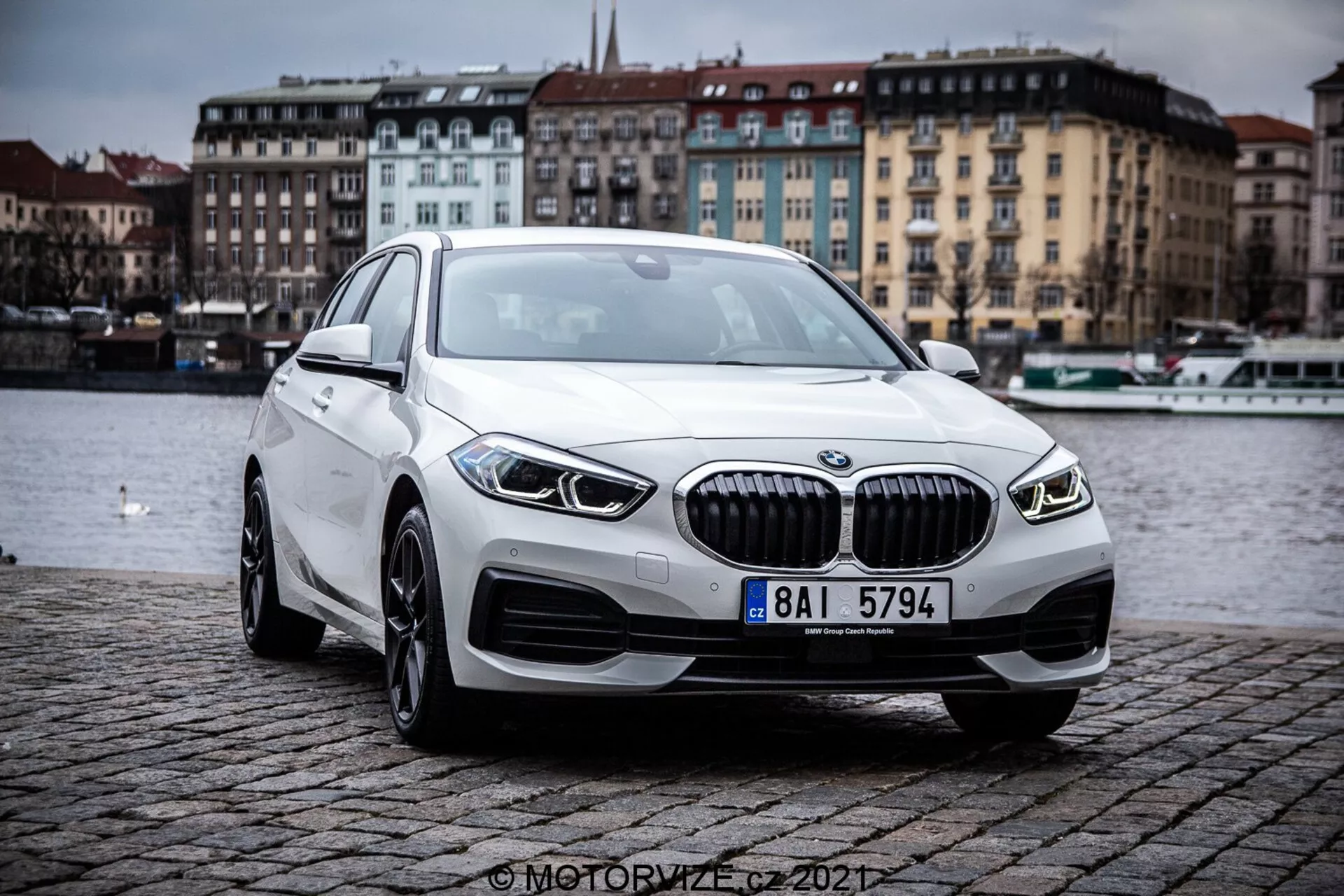 Bild som visar en BMW 1er-Reihe 2019 (BMW 116i) frånt en trekvartsperspektiv framifrån, vilket framhäver den moderna lyxsedanens konturer och designelement. Bilen är placerad utomhus på en kullerstensbank nära en vattensamling under en molnig himmel. Framträdande drag inkluderar en framgrill med vertikala lameller, eleganta LED-strålkastare, integrerade dimljus, lättmetallfälgar och en aerodynamisk profil.