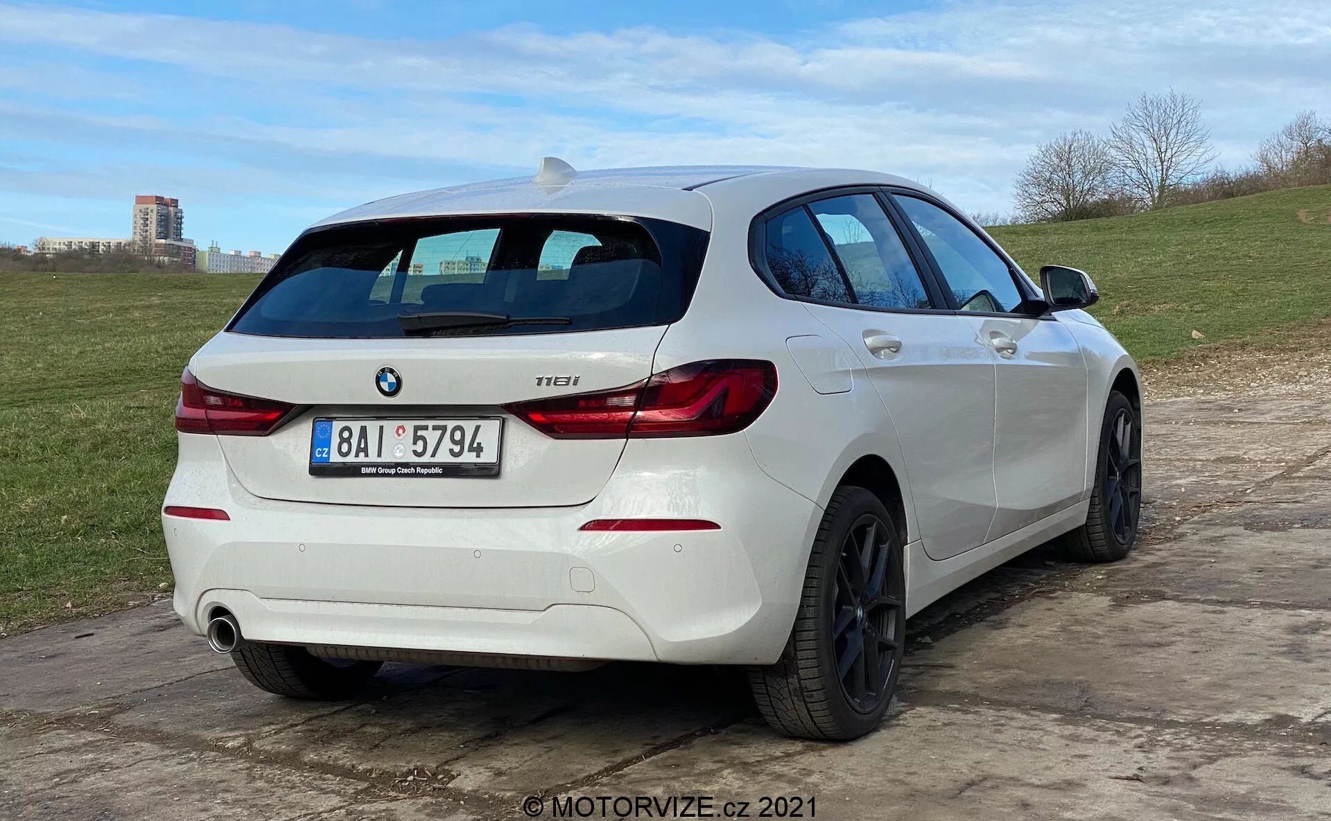 En trekvartsvy bakifrån av en vit BMW 1-serie 118i halvkombi, parkerad på ojämn mark. Bilen har karakteristiska baklyktor i 'L'-form, en BMW-märke och en modellbeteckning som lyder '118i'. Den har ett dubbelt avgasrör med ett rör synligt på högra sidan av bakre stötfångaren. Fordonet är utrustat med svarta flerspoksaluminiumfälgar och en bakre vindrutetorkare. Ett takspoiler bidrar till det sportiga utseendet och sidprofilen avslöjar något tonade fönster, veck i karossen samt dörrhandtag och sidobackspeglar som matchar bilens färgschema. Den låga kameravinkeln betonar bilens hållning och hjuldesign.