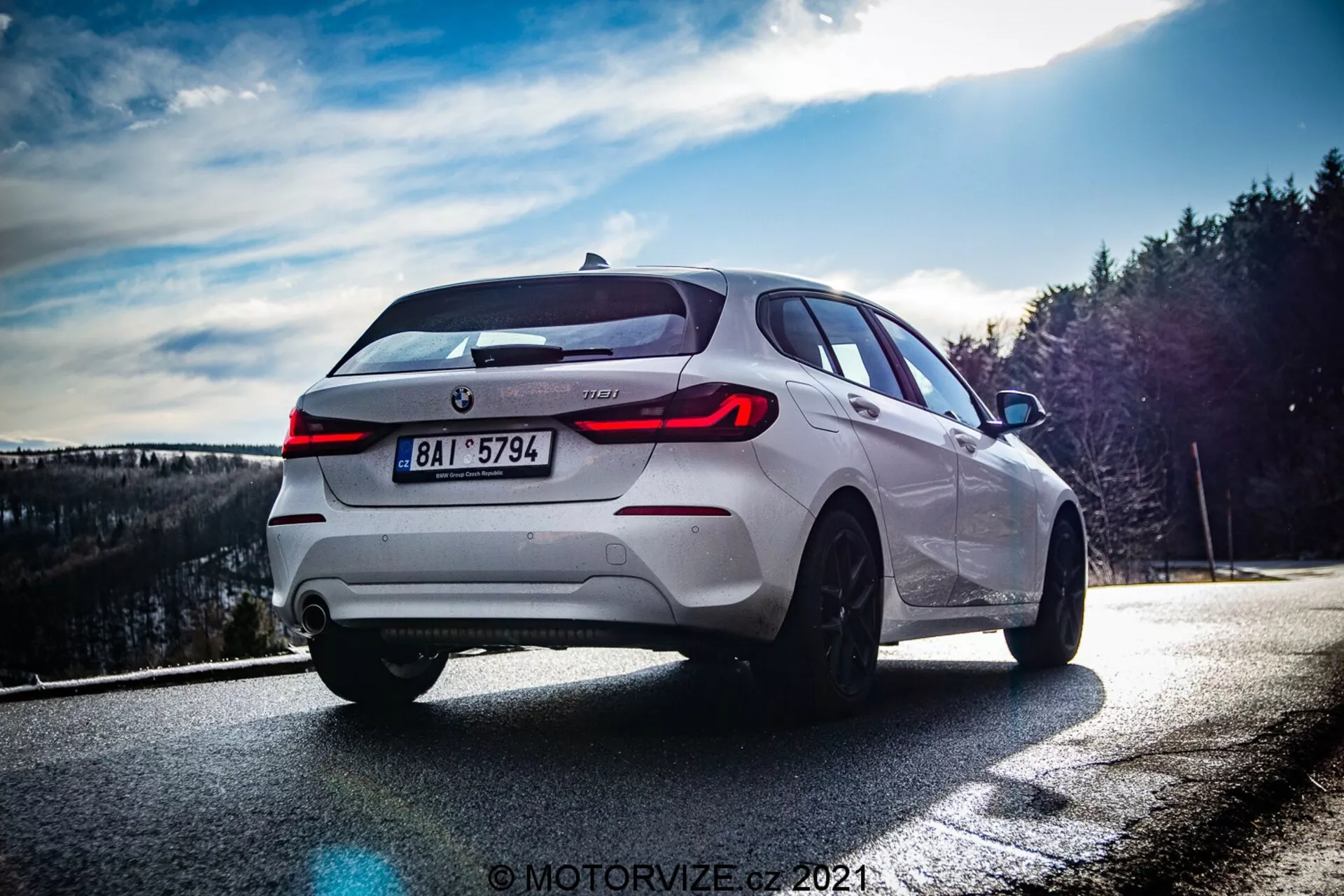 Bilden visar en vit BMW 1-serie 2019 (BMW-116i) fotograferad från en låg och något bakre trekvartsvinkel, vilket betonar bilens högra baksida. Dagsljusinställningen, troligtvis sent på eftermiddagen, skapar dynamiska kontraster som framhäver designlinjerna. Framträdande inslag inkluderar distinkta LED-bakljus, centralt placerad tillverkaremblem och modellbeteckning, dubbelt avgasrörssystem, flerspåriga fälgar i legering, lågprofildäck, synliga skivbromsar och en sportig halvkombisiluett med sluttande bakruta och takspoiler. Fotots komposition och ljuseffekter tillför en dramatisk touch, med suddig bakgrund för att fokusera enbart på fordonet.