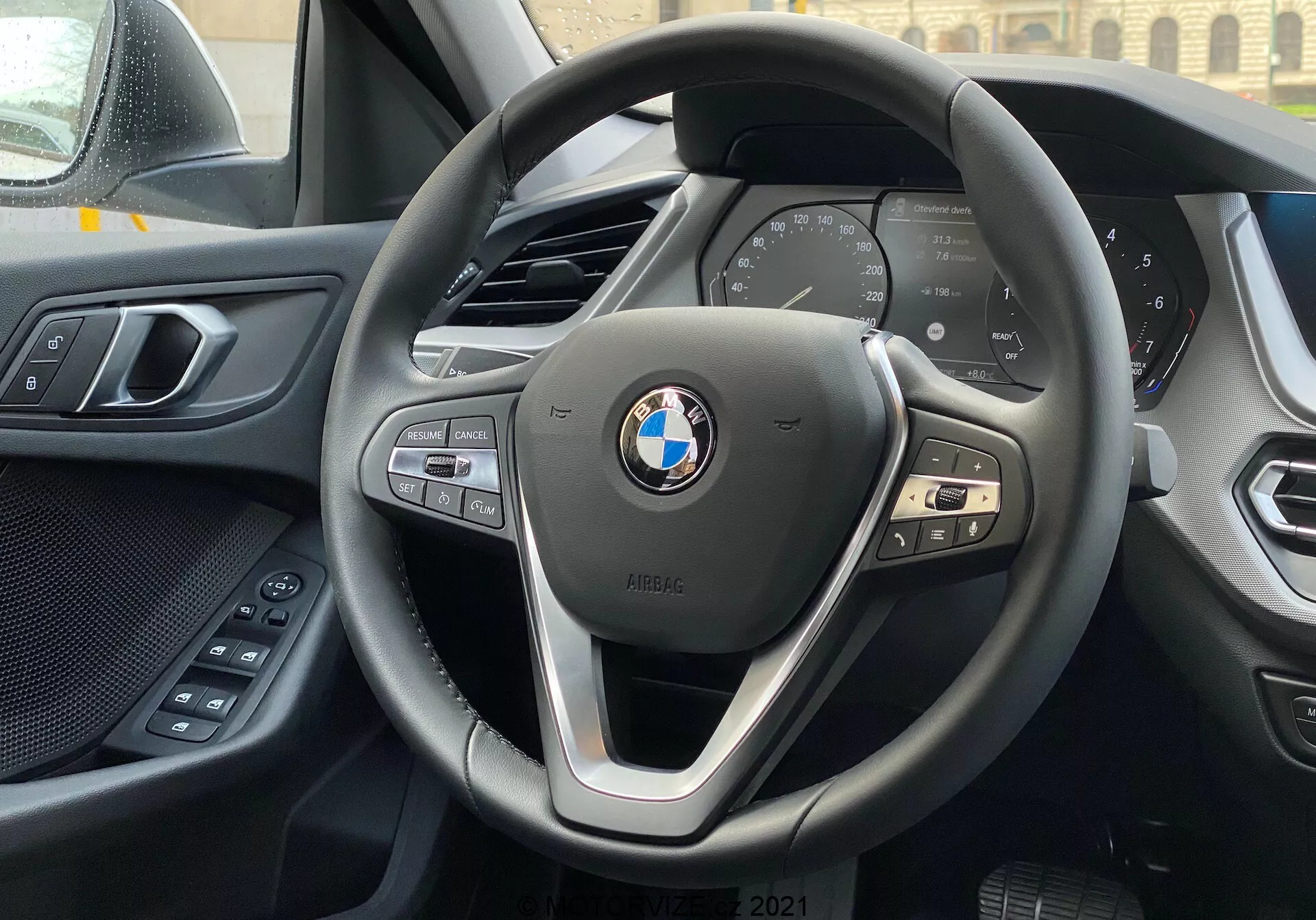 Interiörvy av en BMW 1-serie 2019 (BMW 116i) från förarsätet med fokus på den moderna treekrade BMW-ratten med kontroller för multimedia, telefon och farthållare, och den digitala instrumentpanelen som visar viktiga fordonsdata som hastighet och varvtal. Den övre kanten av mittkonsolen är synlig i bakgrunden och till vänster syns en del av förardörren med elektriska bekvämlighetsreglage. Instrumentbrädan och dörrpanelen visar en kombination av mjuka material och högkvalitativt hantverk. BMW-emblemet och iPERFORMANCE-märket är tydligt synliga på ratten, vilket tyder på effektivitet eller hybridteknik.