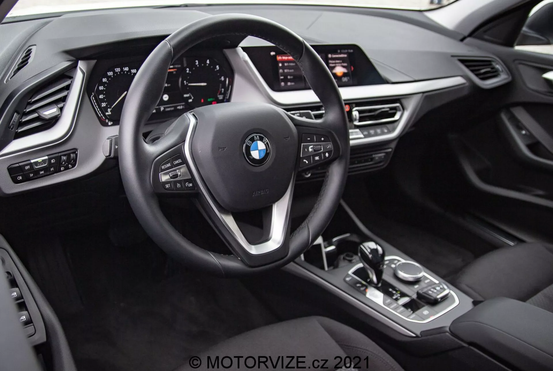 Interiörvy av BMW 1er-reihe 2019 (BMW 116i) från förarens perspektiv. Bilden framhäver BMW-emblemet på den treekrade ratten med multifunktionsknappar och växelpaddlar. Bakom ratten finns en helt digital instrumentpanel med upplysta mätartavlor och olika informationsdisplayer. Mittkonsolen har en växelväljare för automatlåda, iDrive-kontrollvred, knappar för körlägen och en stor infotainmentskärm vinklad mot föraren. Interiördesignen innehåller läderliknande texturer, metalliska accenter och ytterligare kontroller för strålkastare, fönster och spegelinställningar med minnesfunktioner för förarsätet.