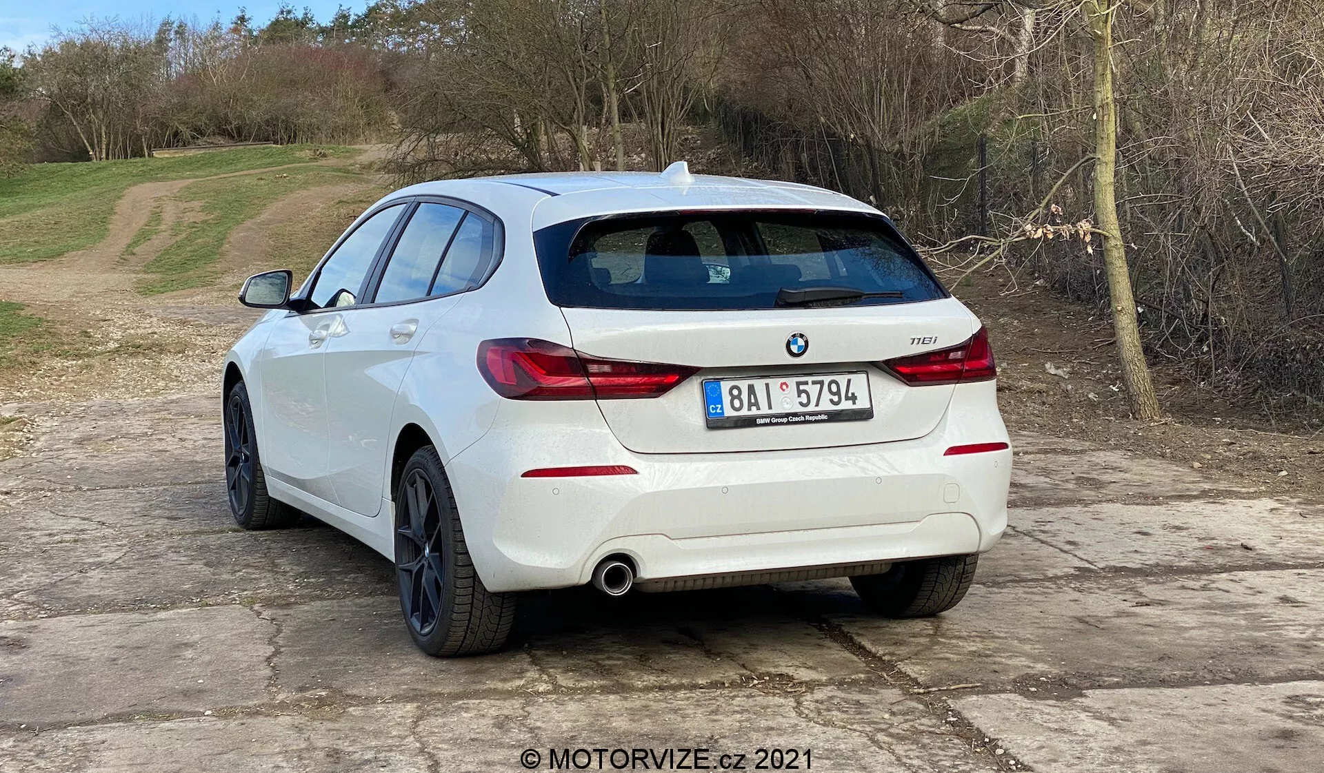 Bilden visar en vit BMW 1er-reihe 2019 (BMW 116i) hatchback sedd från en trekvartsvinkel bakifrån. Fordonets bakre del, inklusive horisontellt orienterade bakljus, dubbla avgasrör och parkeringssensorer integrerade i bakre stödfångaren, är tydligt synliga. BMW-emblemet och modellmärket är synliga på den bakre luckan. Bilen är parkerad på en grusig yta med en naturlig, lantlig bakgrund. Bilden antyder bilteman som fordonsdesign, samtida bilmodeller, BMW-specifik innehåll, avgasystem och parkeringsassistans teknologi. Förekomsten av en MOTORVIZE.cz 2021 vattenstämpel indikerar ett ursprung i en bilrecension eller publikation.