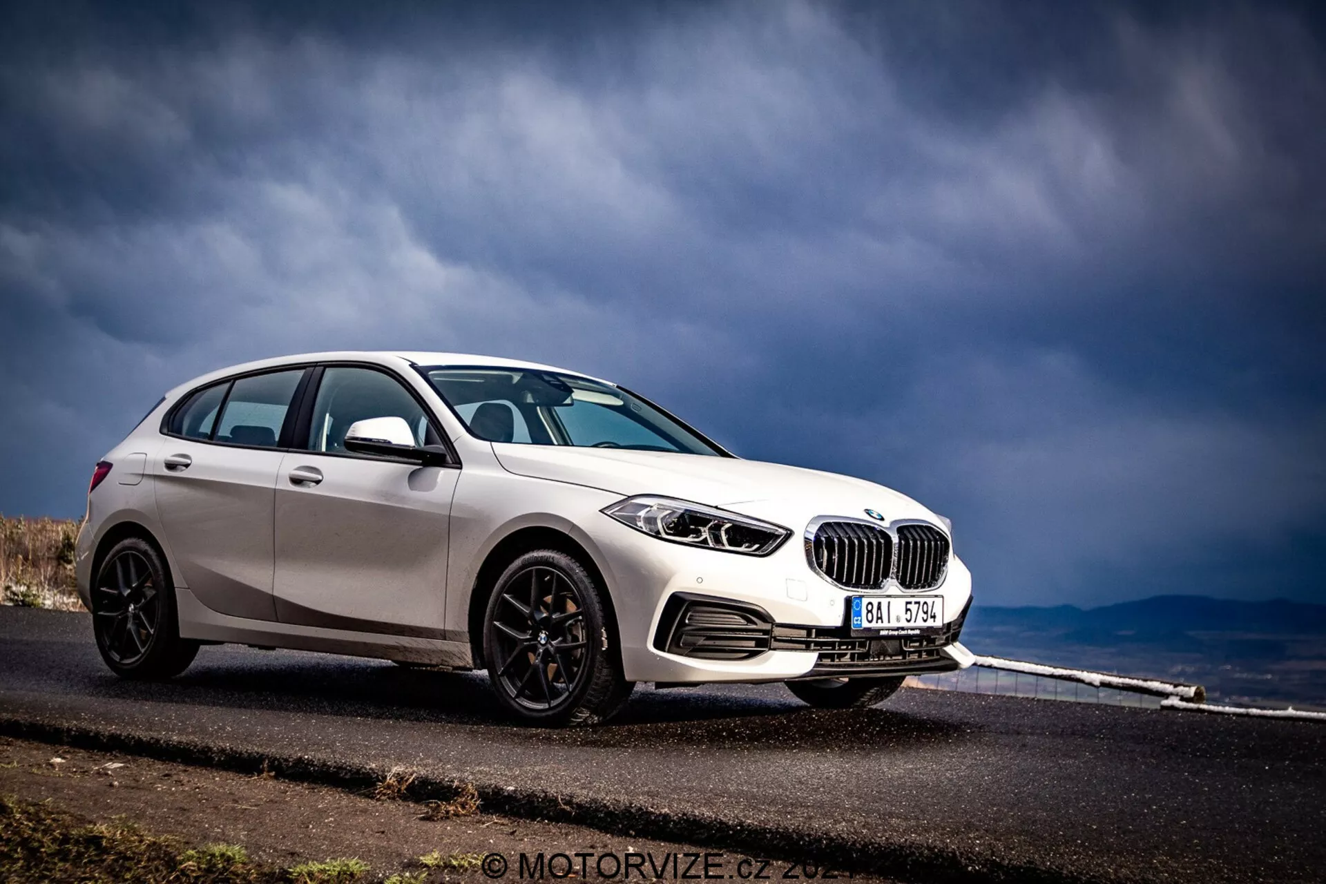 BMW 1-serien 2019 (BMW-116i) fångad i en dramatisk trekvarts frontvy, visar dess främre och sidoprofiler. Bilen, färdig i ljus vit med kontrasterande svarta accenter som spegelskal, fönsterlist och flerspäkiga hjul, är placerad på vänster sida av ramen mot en bakgrund av en asfalterad vägen som sträcker sig i fjärran och stormigt himmel. Synliga funktioner inkluderar detaljerade strålkastare med cirkulära element, framträdande njurgrill, främre stötfångare med luftintagsventiler och potentiella parkeringssensorer.