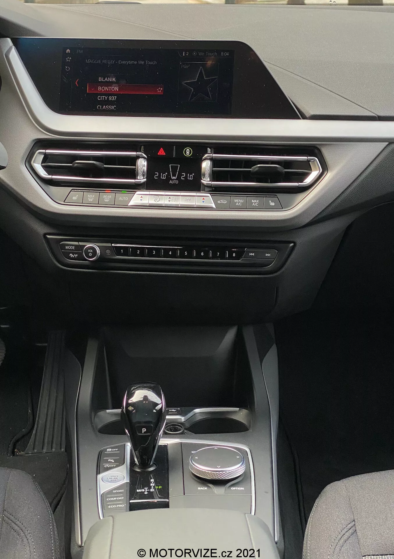 Interiörvy av en BMW 1er-reihe 2019 (BMW-116i) som visar en modern och lyxig kabin med fokus på mittkonsolen och instrumentbrädan. Perspektivet är från baksätet, framåt mot infotainment-skärmen som är monterad högst upp på instrumentbrädan. Skärmen visar alternativ för radio, media och bilinställningar. Nedanför skärmen finns horisontellt anpassade luftventiler med metalldetaljer och en dekorativ kant som sträcker sig över hela instrumentbrädans bredd. Nedanför ventilerna syns fysiska knappar och reglage för klimatkontroll och andra inställningar. Automatväxelspaken har en blank svart yta med metalldetaljer, placerad bredvid en roterande ratt. Mittkonsolen inkluderar två mugghållare, ett litet förvaringsfack och en silverfärgad dekorationsdetalj, vilket indikerar en välutrustad kabin som kombinerar läder och högkvalitativa syntetiska tyger, lämplig för fordon i mellanklass till premiumklassen med fokus på teknik och komfort.