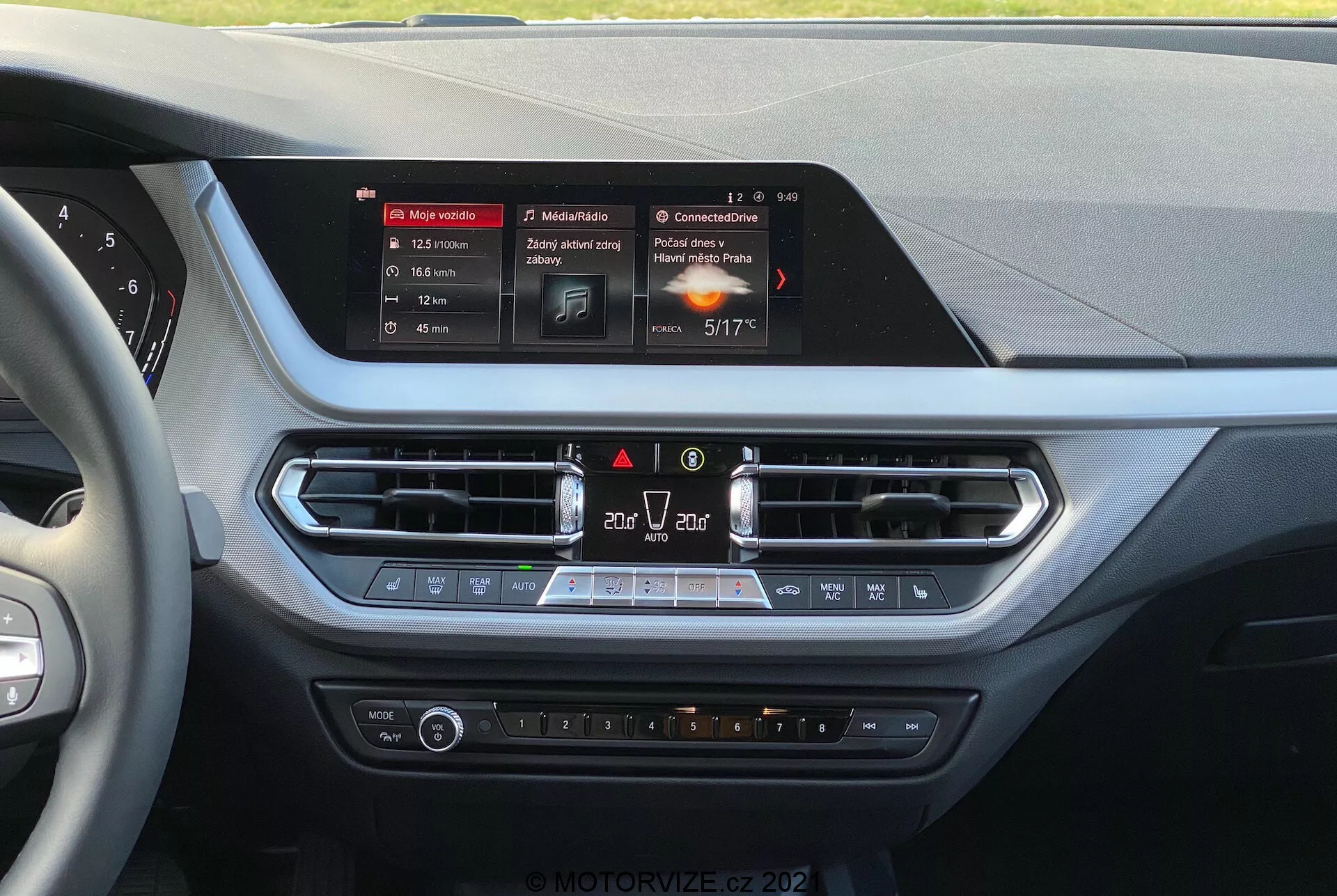 Interiörvy av en BMW 1-serien 2019 (BMW-116i) från förarens perspektiv, som visar mittkonsolen på instrumentbrädan. Centralt finns infotainmentsystemets rektangulära, upplysta pekskärm som visar navigering, media och klimatdetaljer. Under skärmen finns metallklädda luftventiler med nöd- och luftcirkulationskontroller, följt av en klimatkontrollpanel med temperaturvred, fläkthastighet och uppvärmda säten. Materialen har mjuka ytor och kromaccenter, vilket tyder på fokus på förarens och framsätespassagerarens komfort och bekvämlighet.